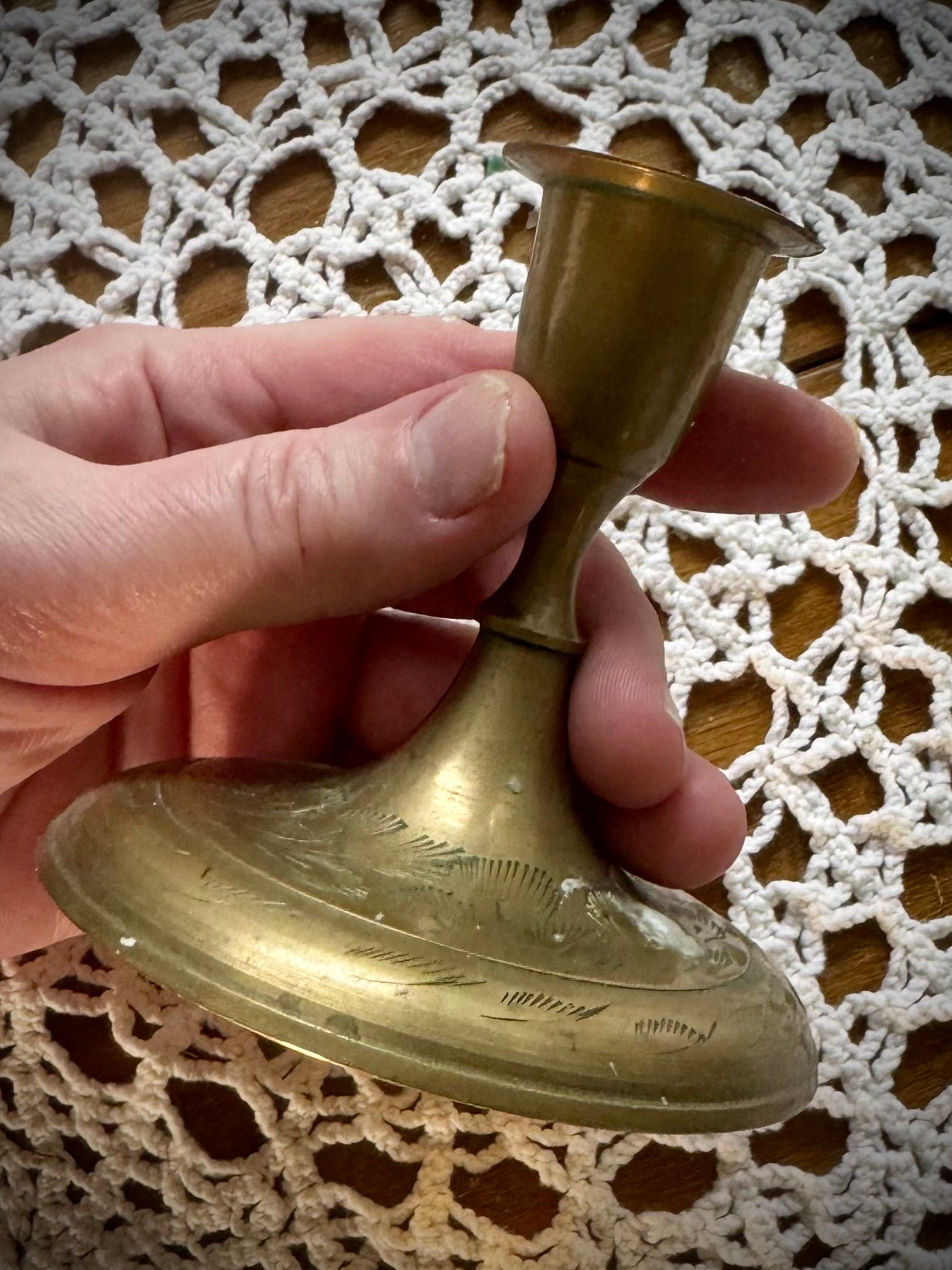 A vintage brass taper candle holder with etching on the base, displayed on a lace tablecloth.