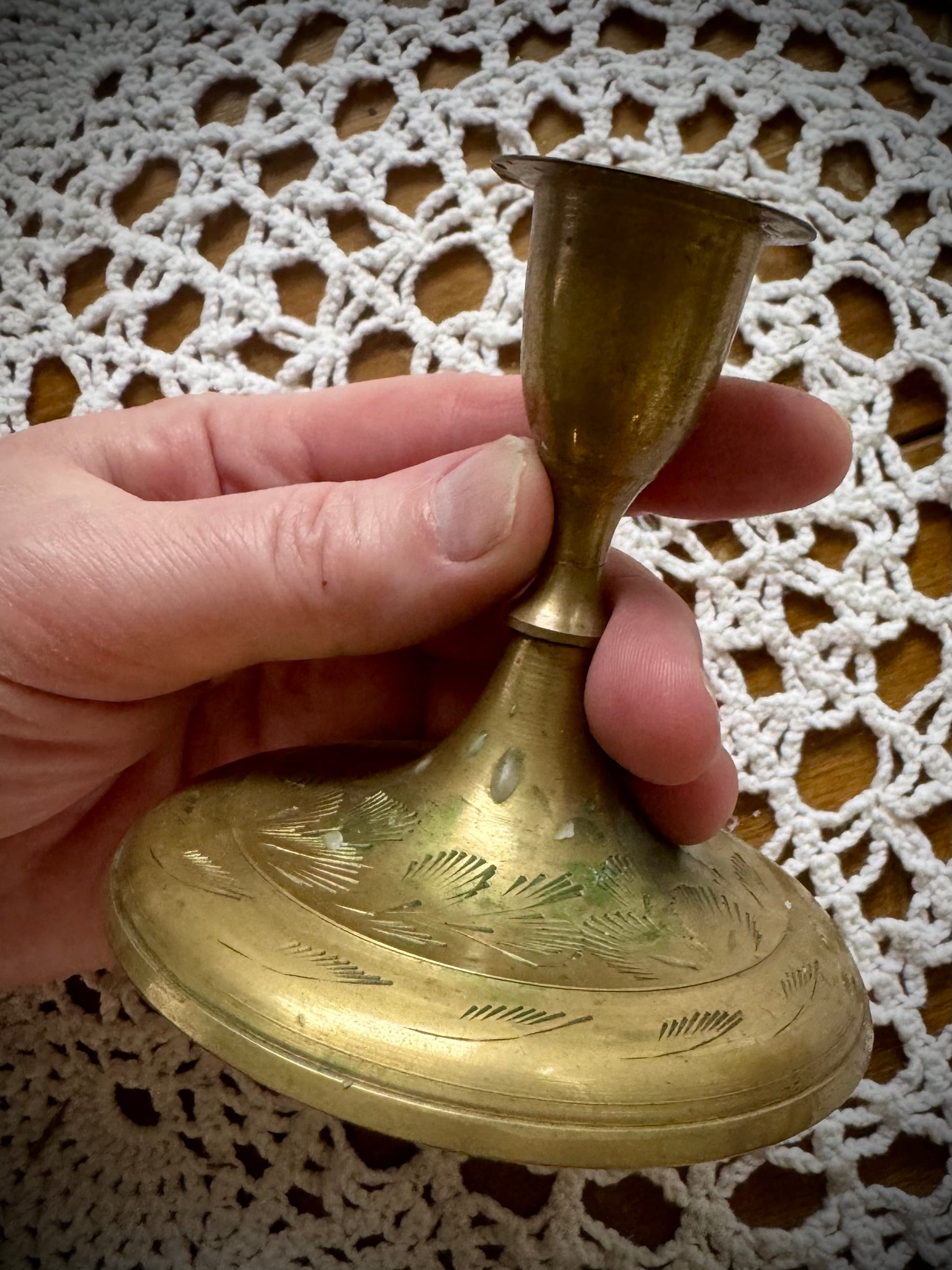 A vintage brass taper candle holder with etching on the base, displayed on a lace tablecloth.