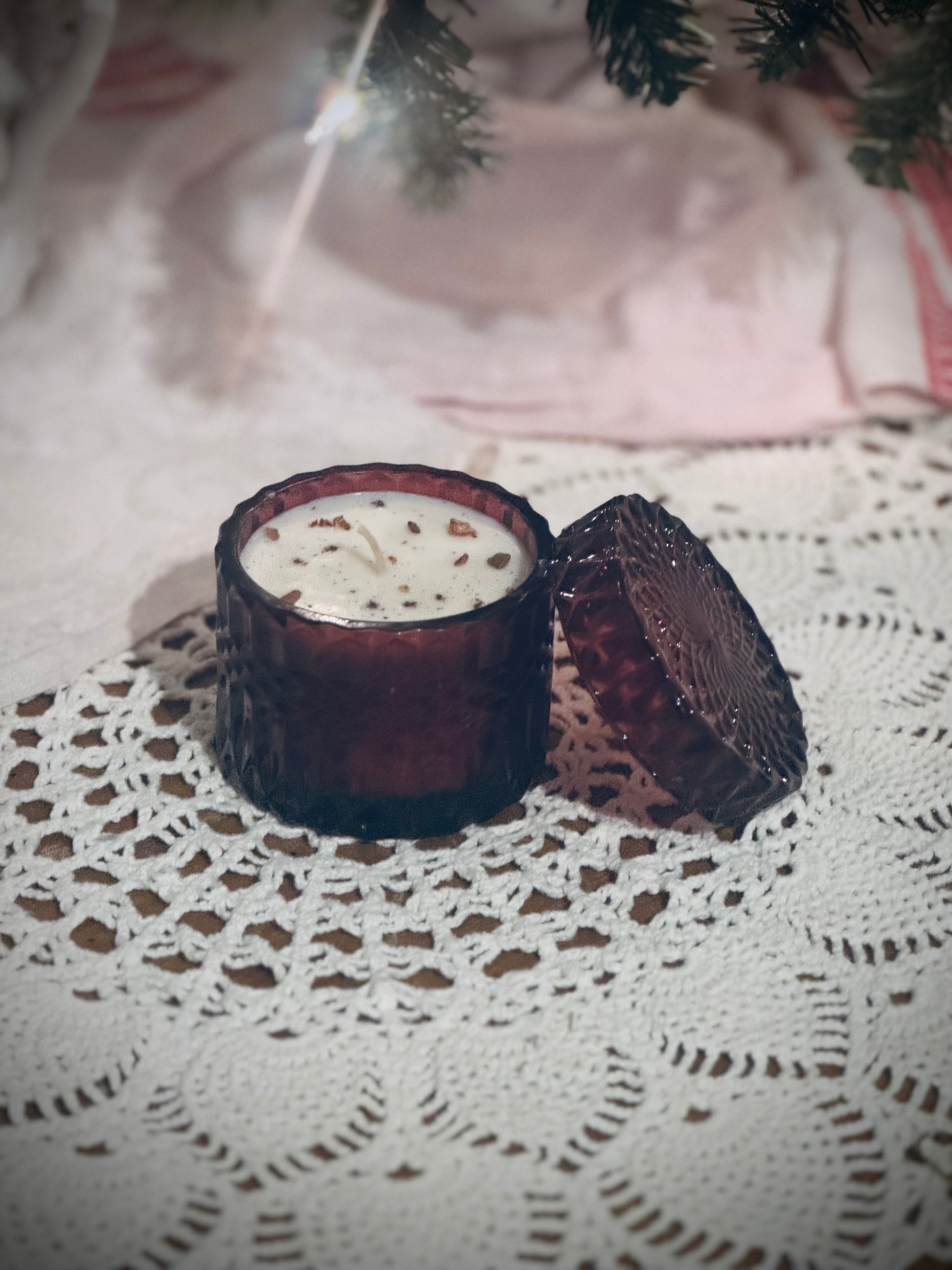 A deep red glass candle vessel with lid placed on a lace tablecloth in front of a decorated Christmas tree.
