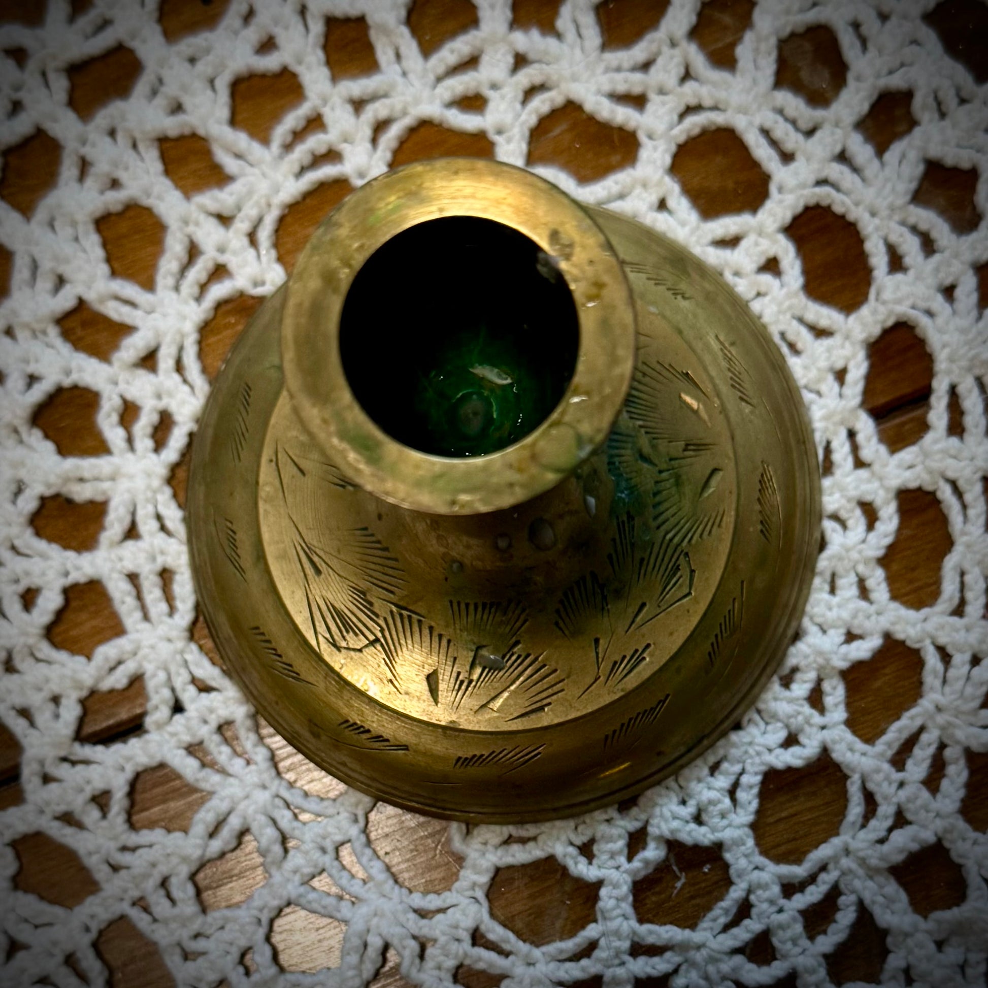 A vintage brass taper candle holder with etching on the base and some green discoloration, displayed on a lace tablecloth.