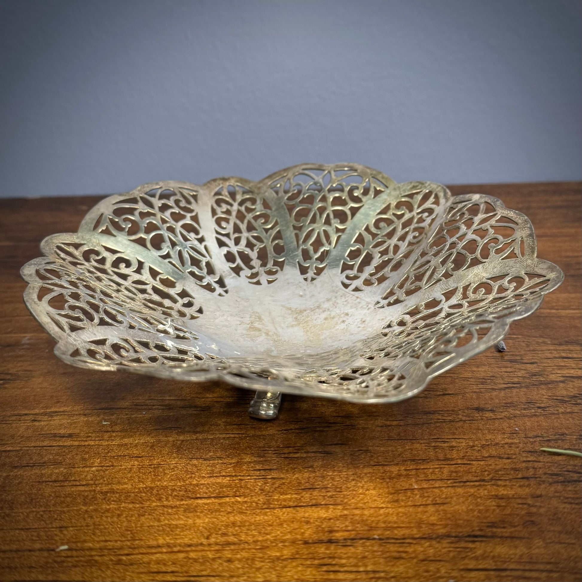 A vintage silver ritual plate with intricate floral lace patterns and three small feet on a wooden surface.