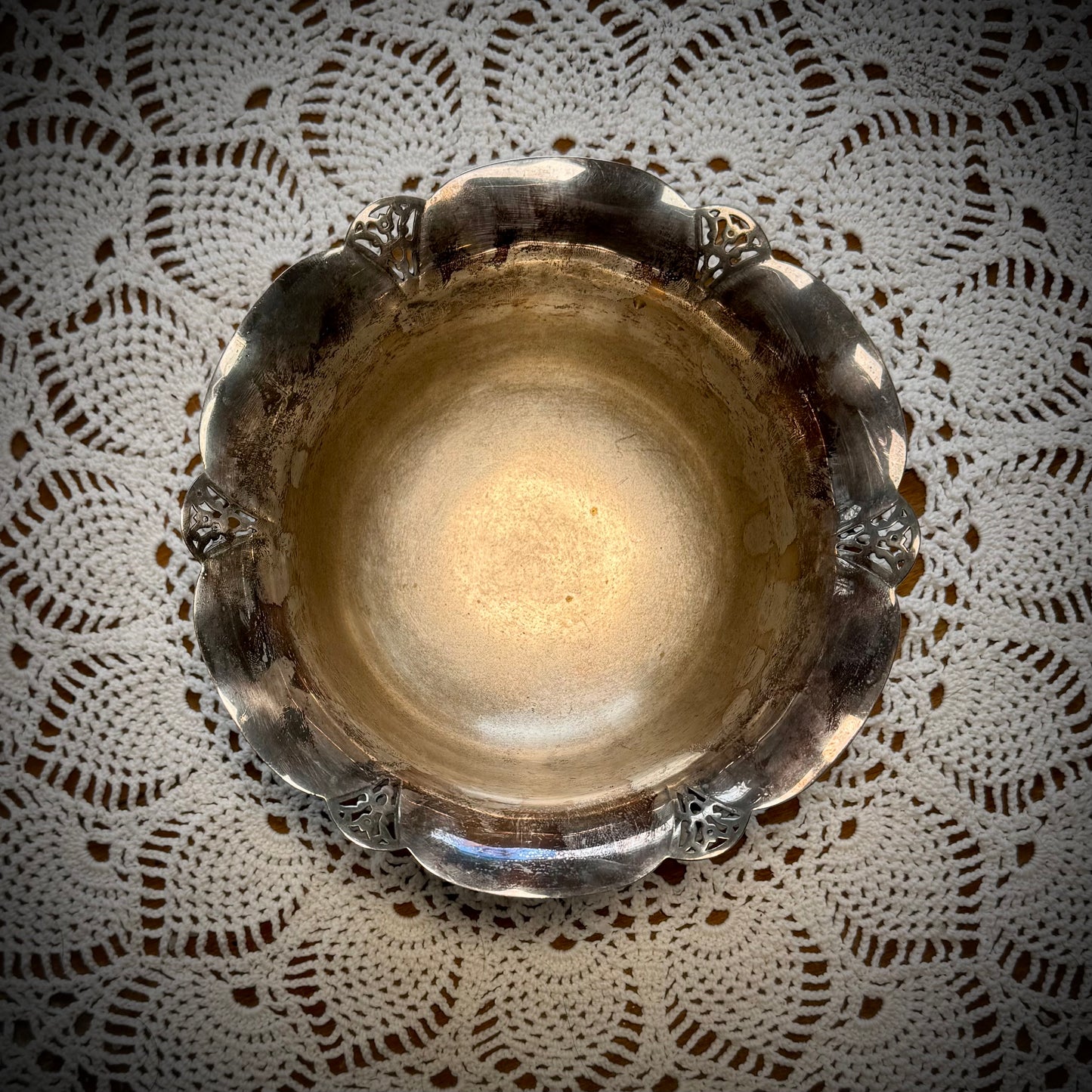 Aerial view of a vintage silver serving bowl with scalloped edges and feet, placed on a table with a lace tablecloth.
