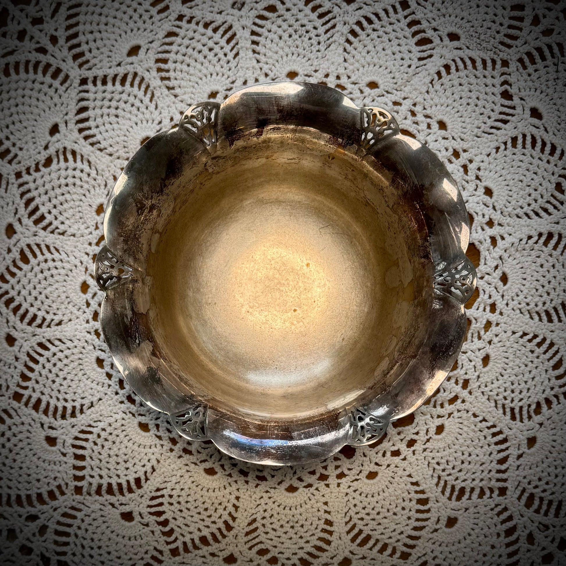 Aerial view of a vintage silver serving bowl with scalloped edges and feet, placed on a table with a lace tablecloth.