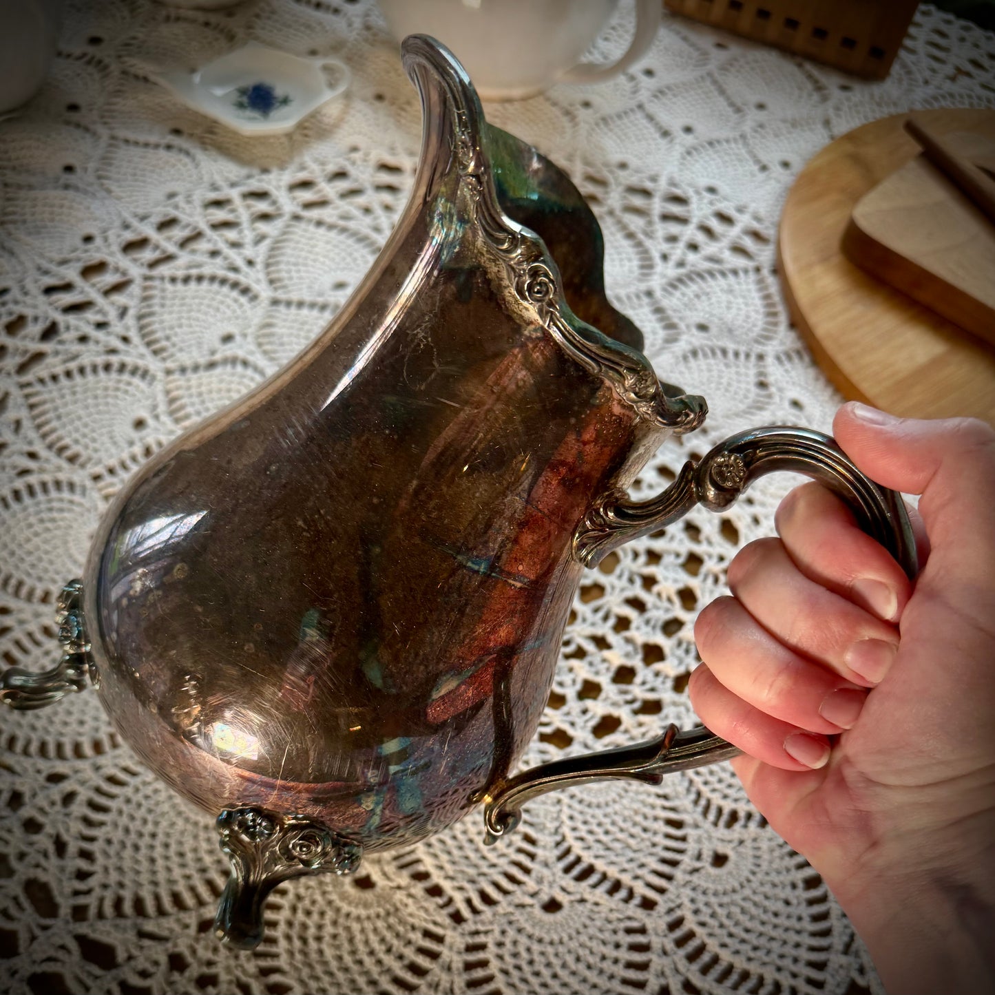 A vintage silver plate water pitcher with floral patterns, featuring a handle and spout, being held by the handle.