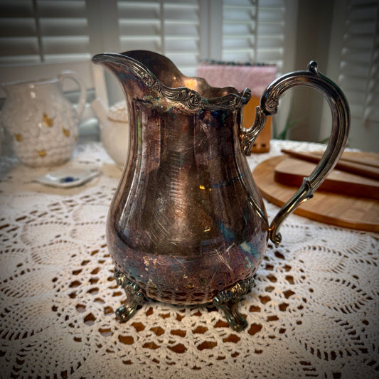 A vintage silver plate water pitcher with floral patterns, featuring a handle and spout, displayed on a table with a lace tablecloth.
