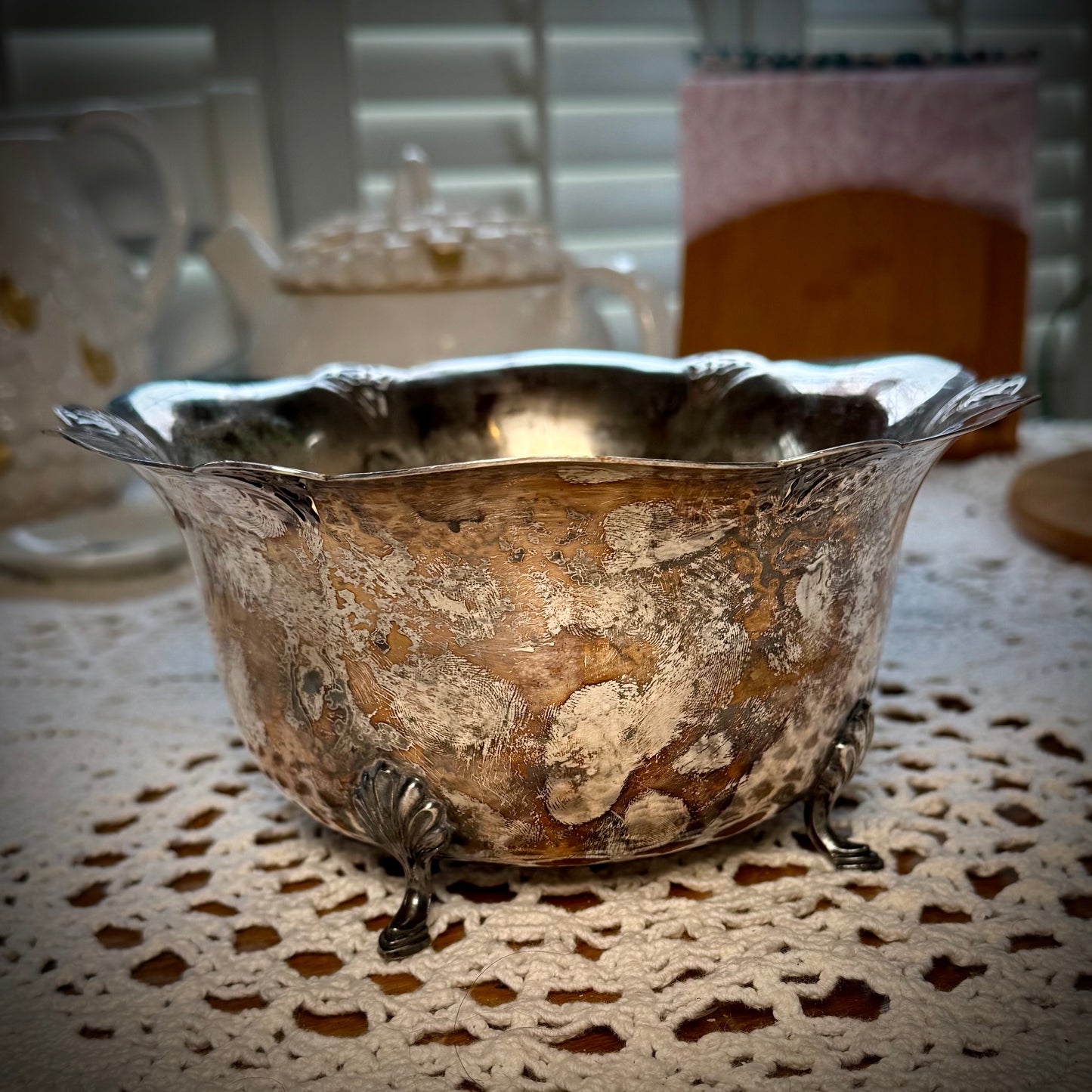 A vintage silver serving bowl with scalloped edges and feet, placed on a table with a lace tablecloth.