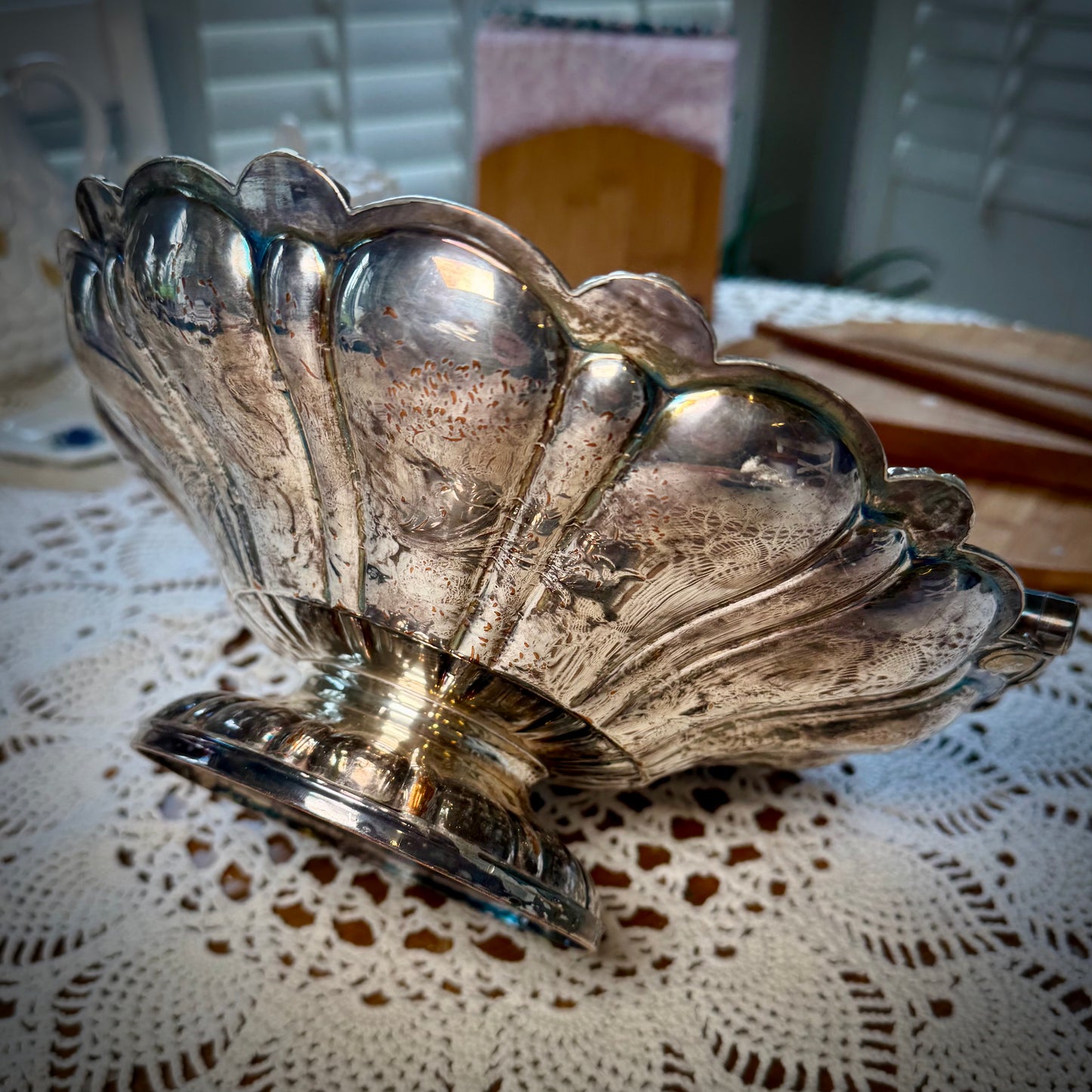 A silver plate basket with a handle, placed on a table with a lace tablecloth, displaying a reflective surface.