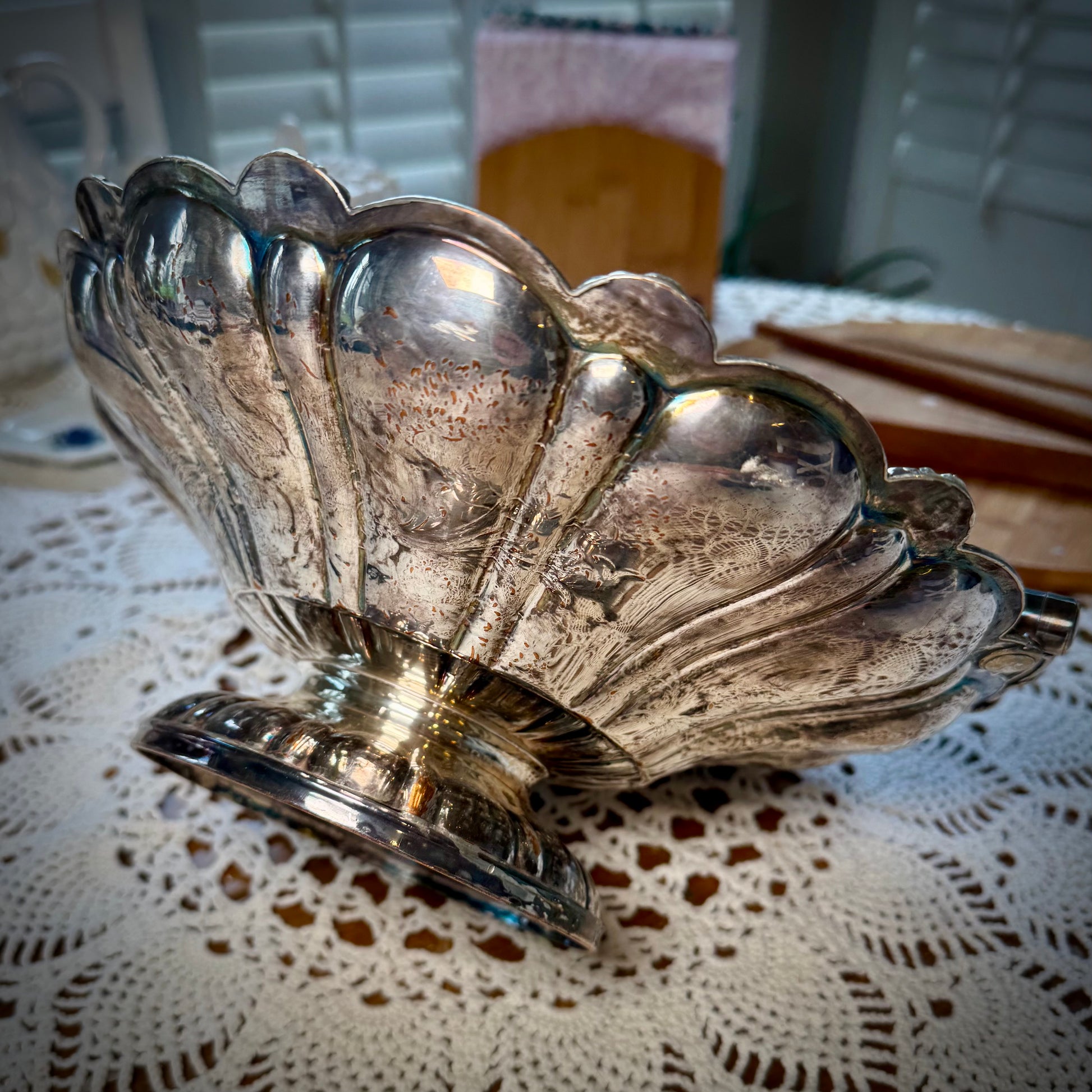A silver plate basket with a handle, placed on a table with a lace tablecloth, displaying a reflective surface.