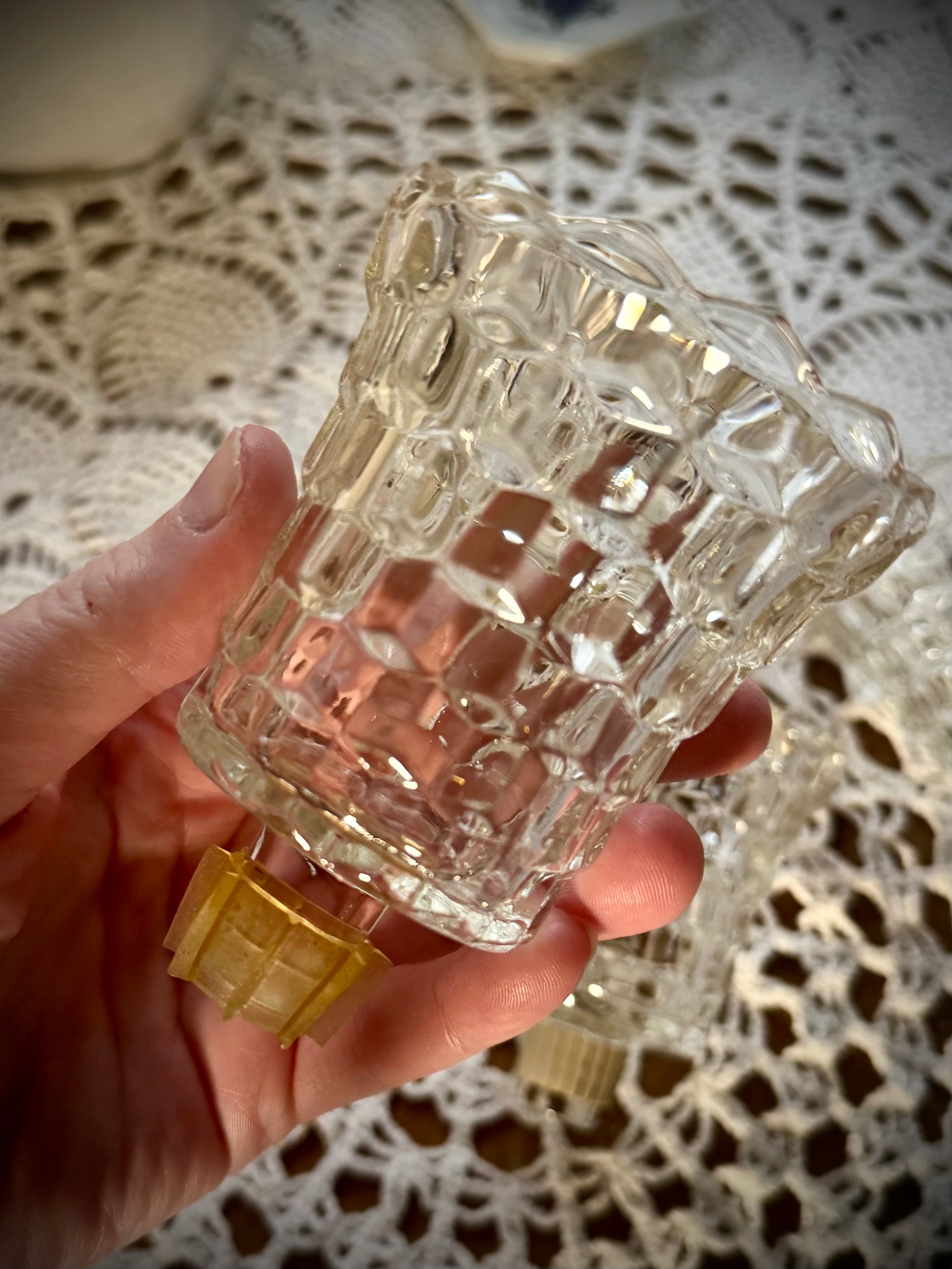 A single glass votive candle holder with a peg and gasket bottom being held above a table with a white lace tablecloth