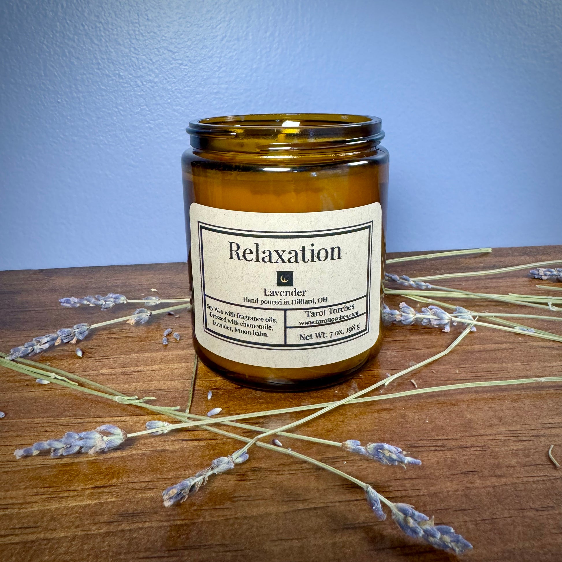 A brown glass candle with a label that reads 'Relaxation' and is infused with lavender fragrance. The candle is placed on a wooden surface with some dried lavender sprigs around it.