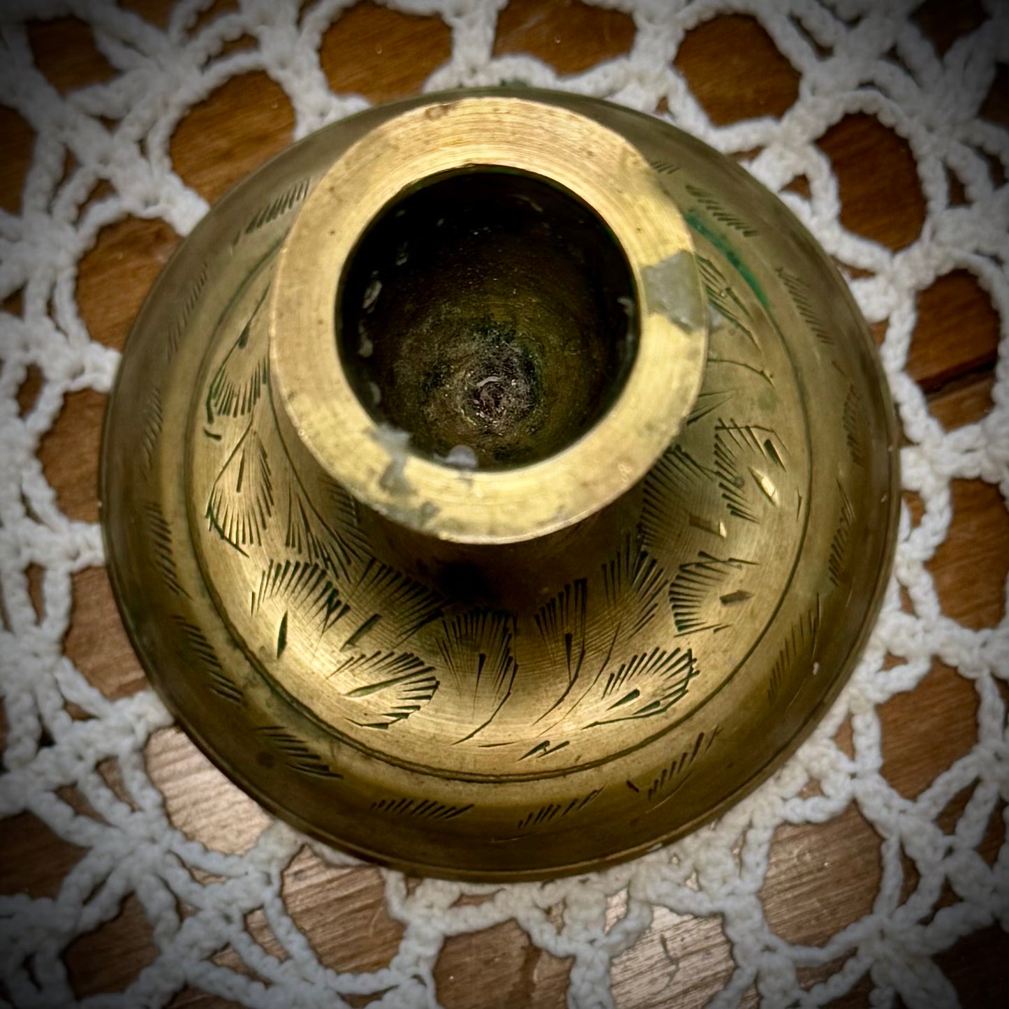 A vintage brass taper candle holder with etching on the base, displayed on a lace tablecloth.