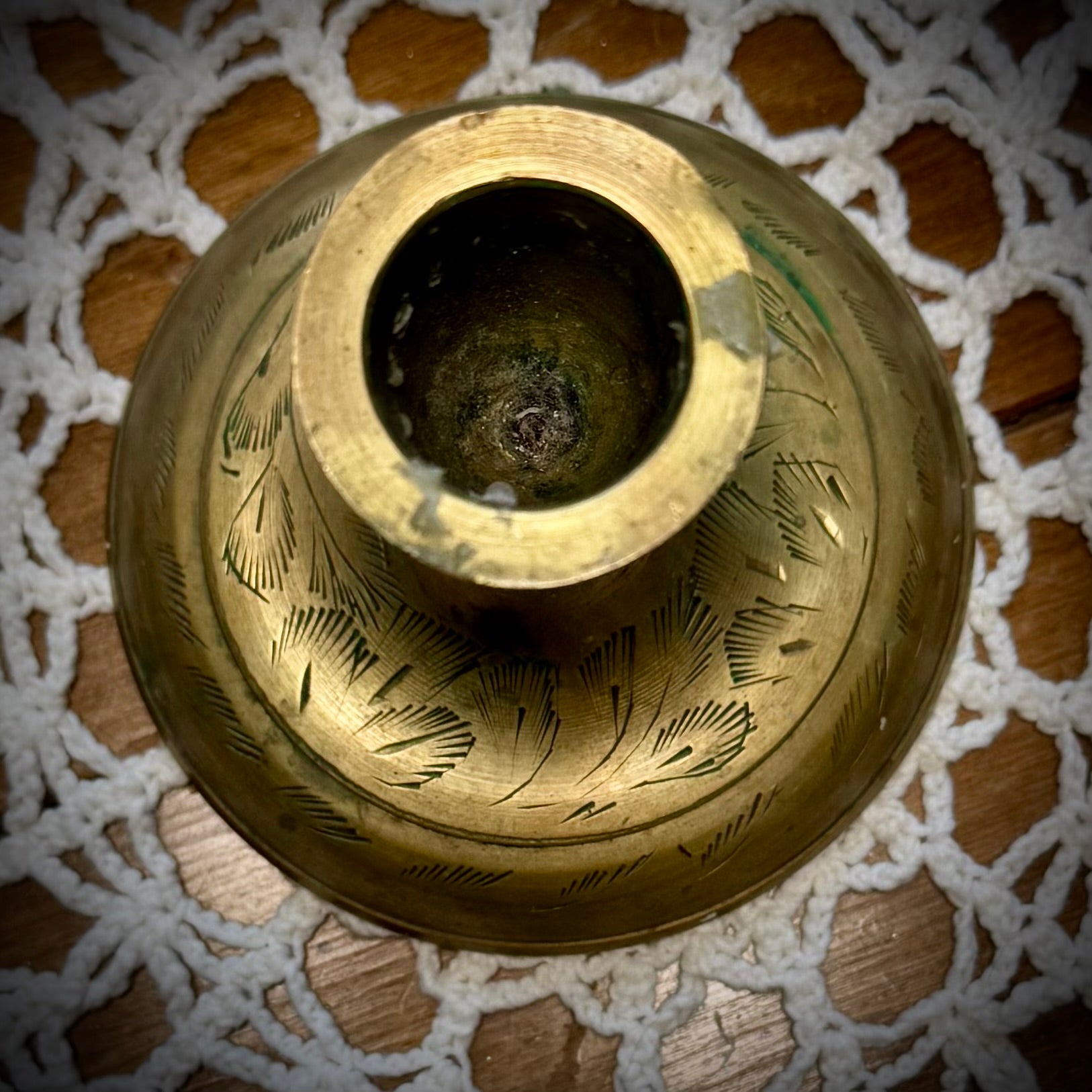 A vintage brass taper candle holder with etching on the base, displayed on a lace tablecloth.