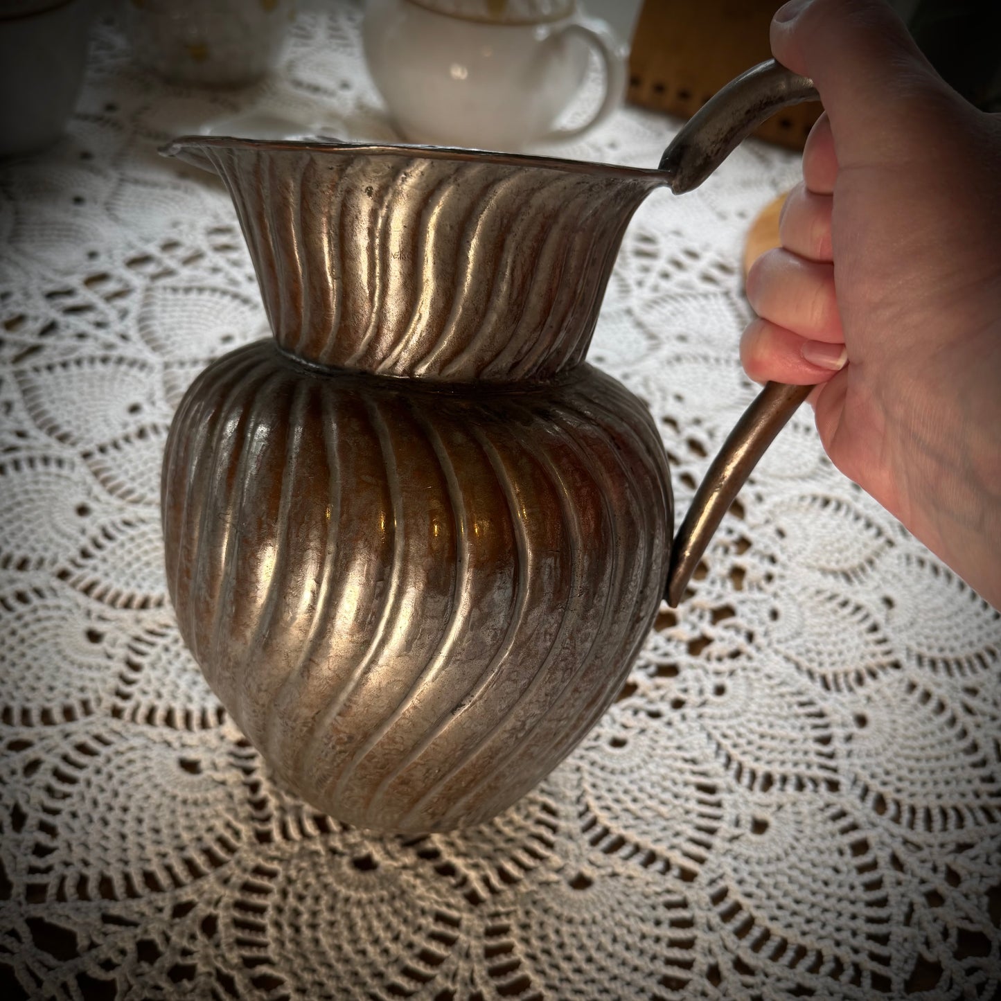 An antique swirl tinned copper pitcher being held up over a table with a white lace tablecloth