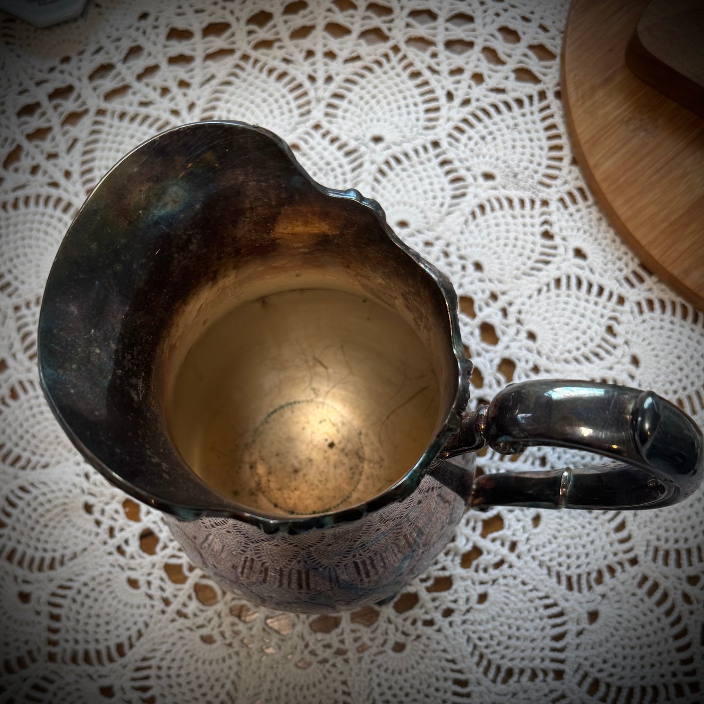 Aerial view of a vintage silver plate water pitcher with floral patterns, featuring a handle and spout, displayed on a table with a lace tablecloth.