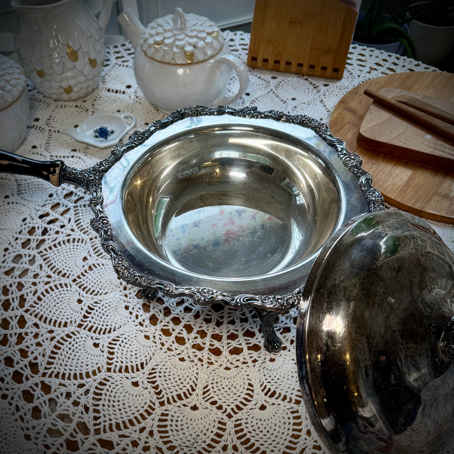 A vintage silver chafing dish with a lid placed next to it, on a table with a lace tablecloth.