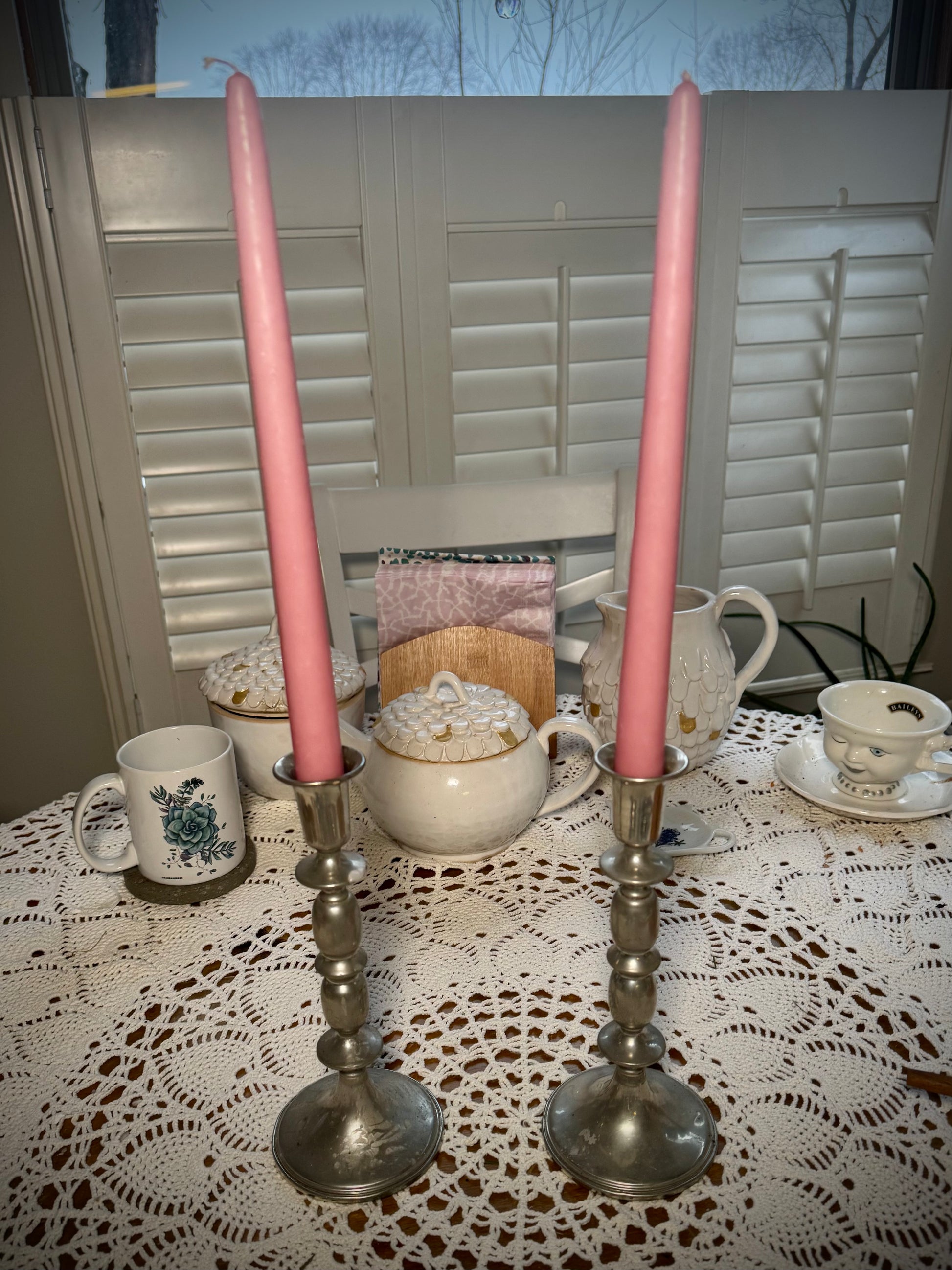 Two tall pewter candlesticks holding tall pink taper candles on a table with a white lace tablecloth and teapot in the background