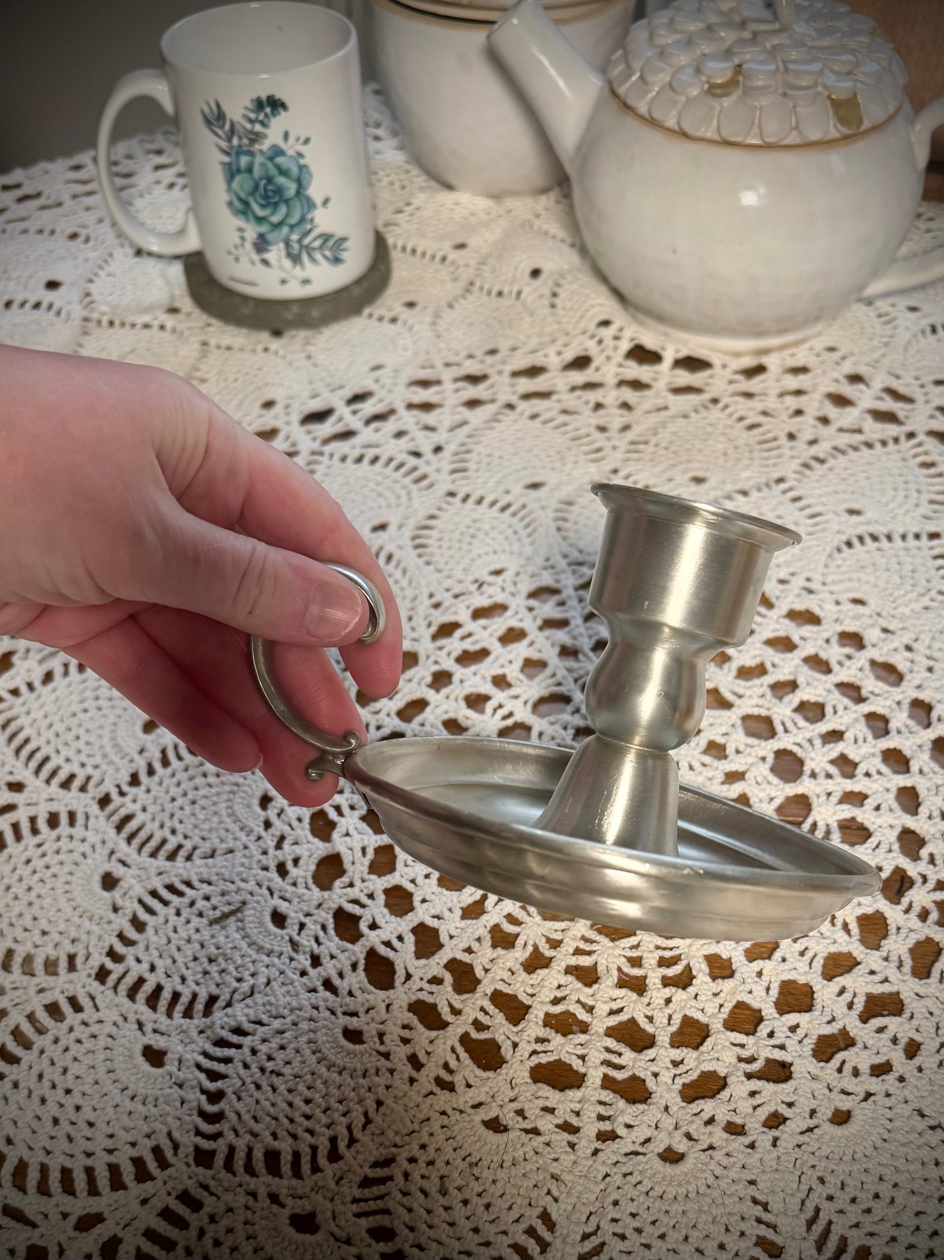 A vintage pewter candlestick with a handle, displayed on a table with a lace tablecloth, against a backdrop of a teapot and cups.
