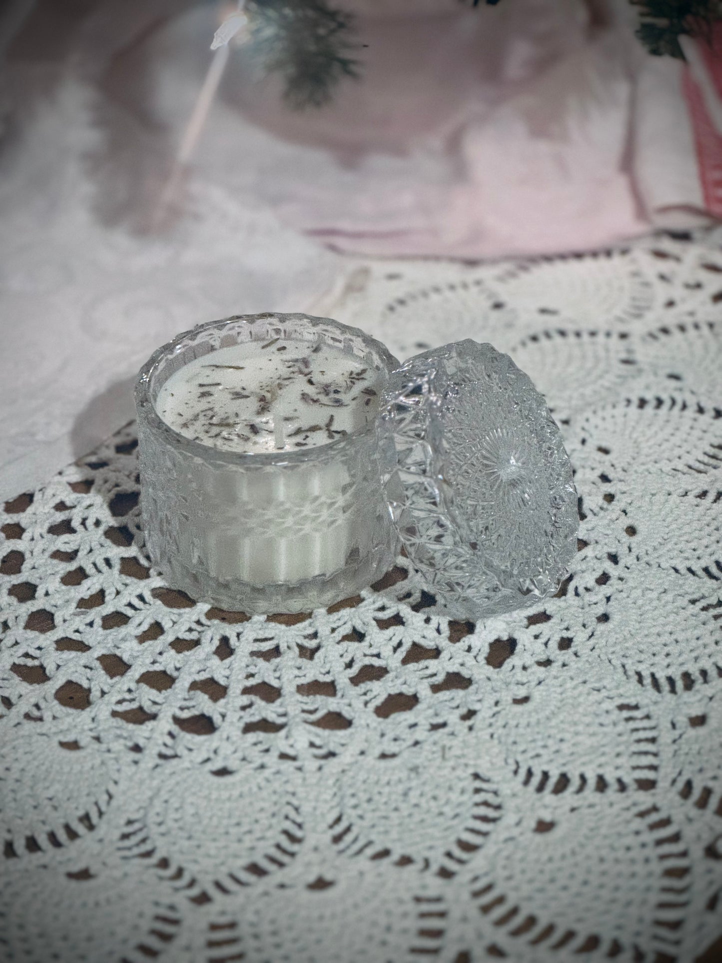 A clear glass candle vessel with lid placed on a lace tablecloth in front of a decorated Christmas tree.