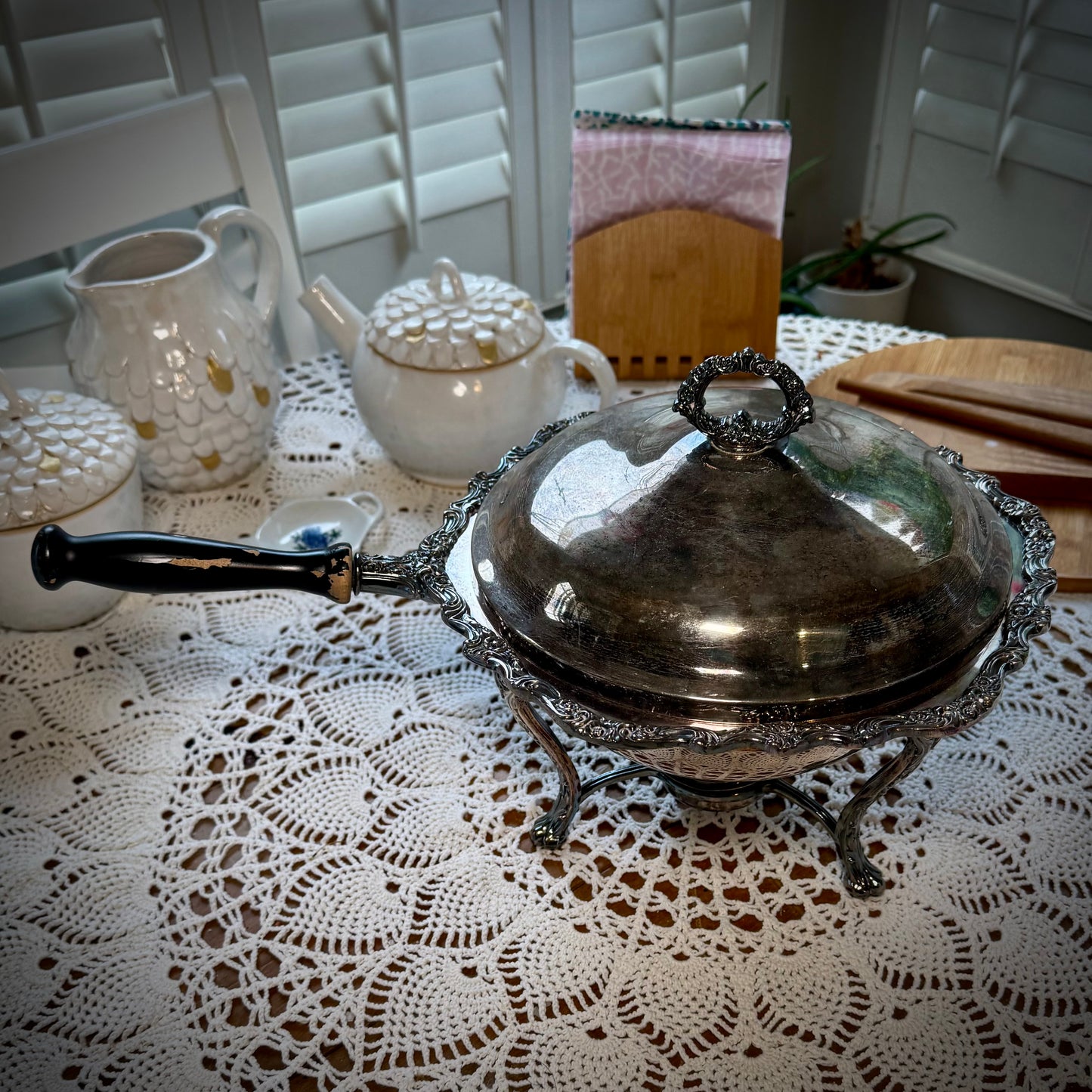 A vintage silver chafing dish with a lid, placed on a table with a lace tablecloth.