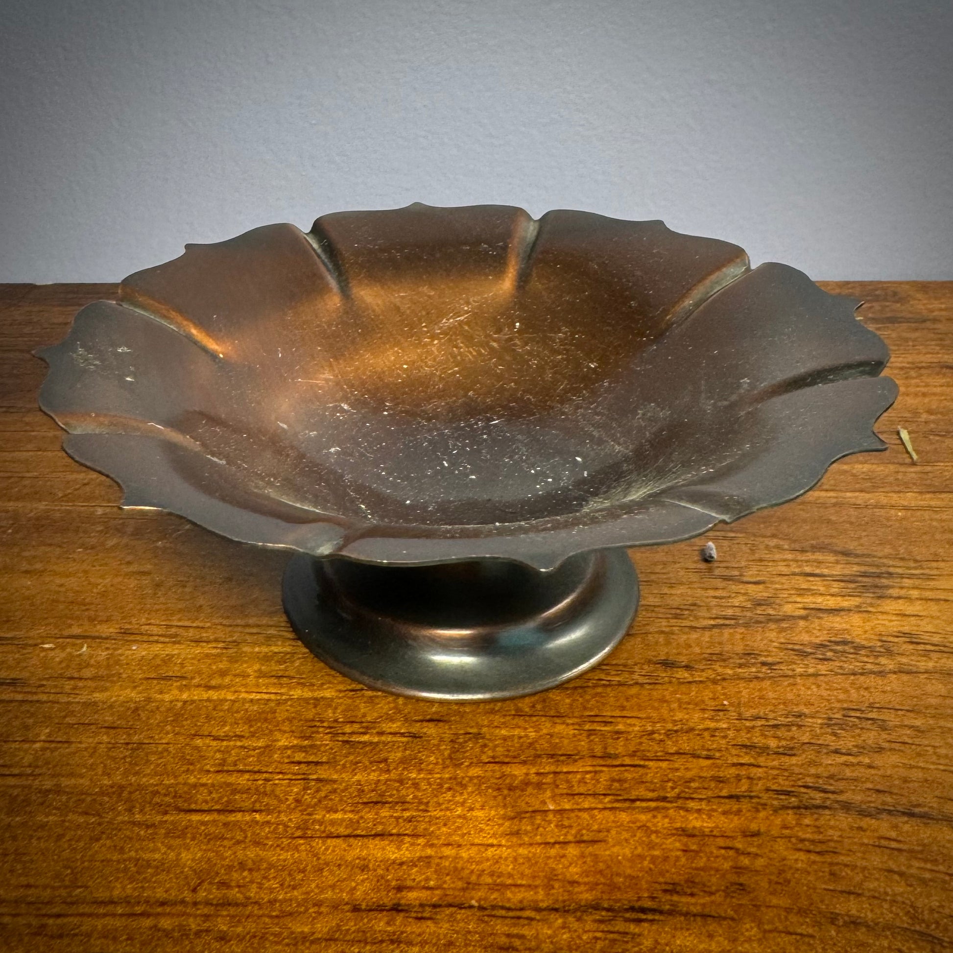 A vintage ritual bowl with a scalloped edge and a deep dark copper color, placed on a short and sturdy stand.