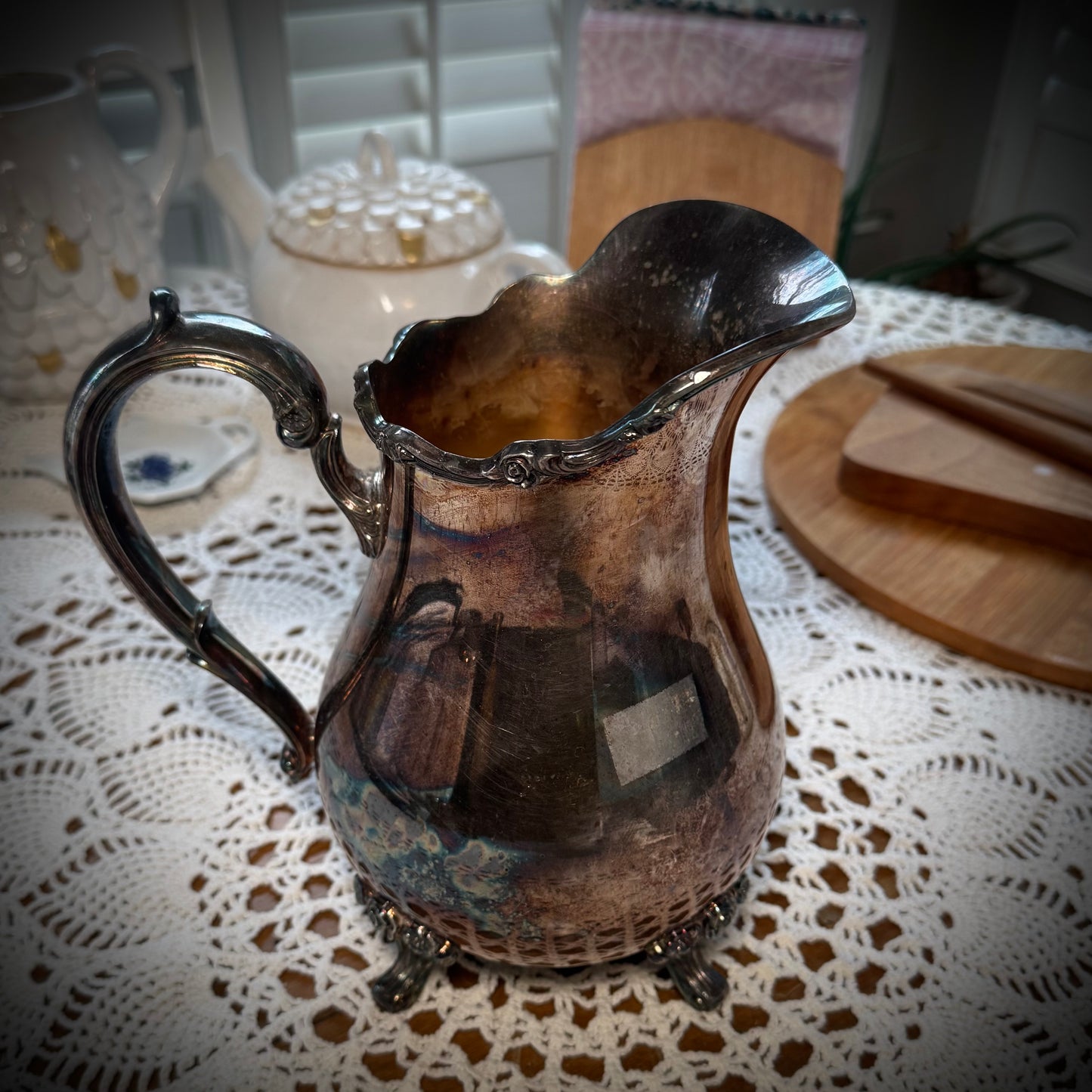 A vintage silver plate water pitcher with floral patterns, featuring a handle and spout, displayed on a table with a lace tablecloth.