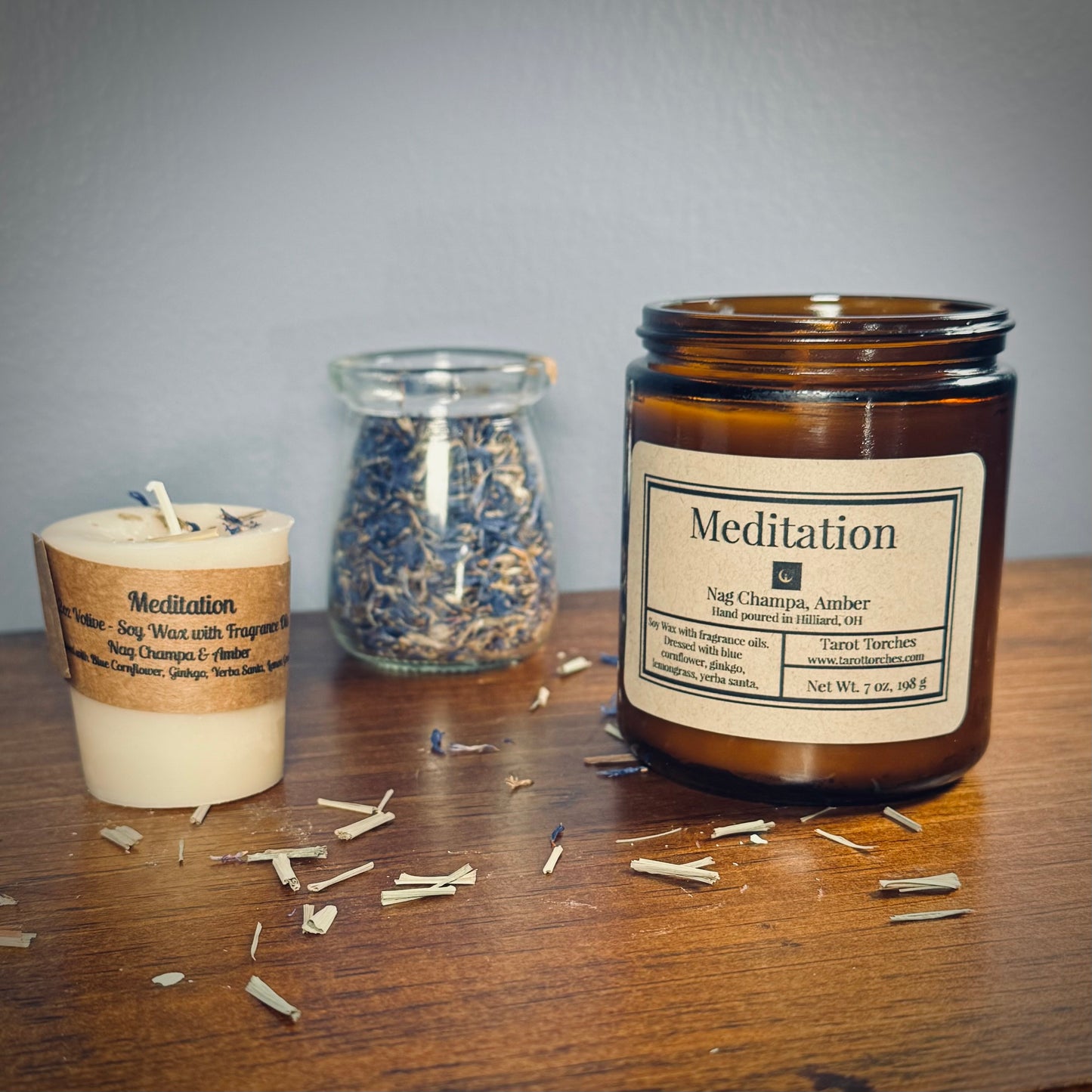 A white votive candle and amber glass candle both reading "Meditation" sit on a wooden surface with a glass jar of herbs between them.