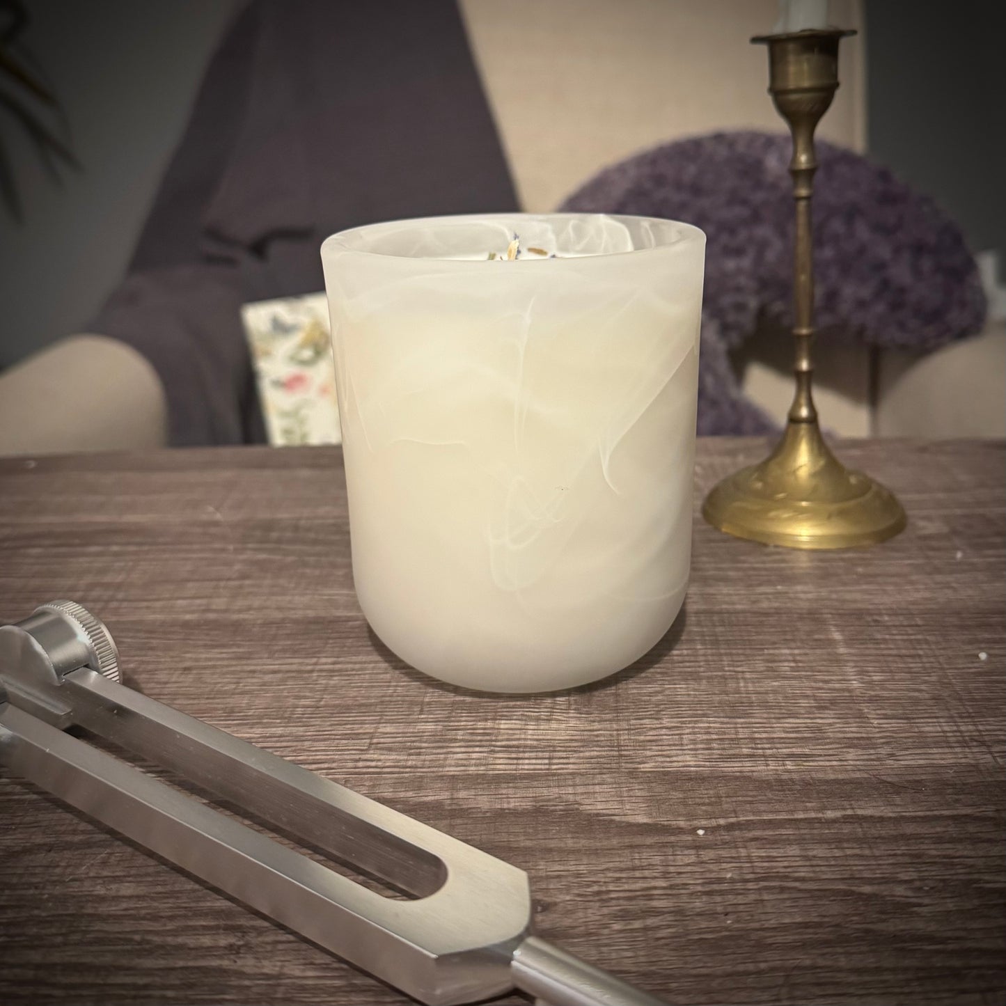 A white swirl glass candle sits on a table behind a tuning fork and in front of a brass candlestick