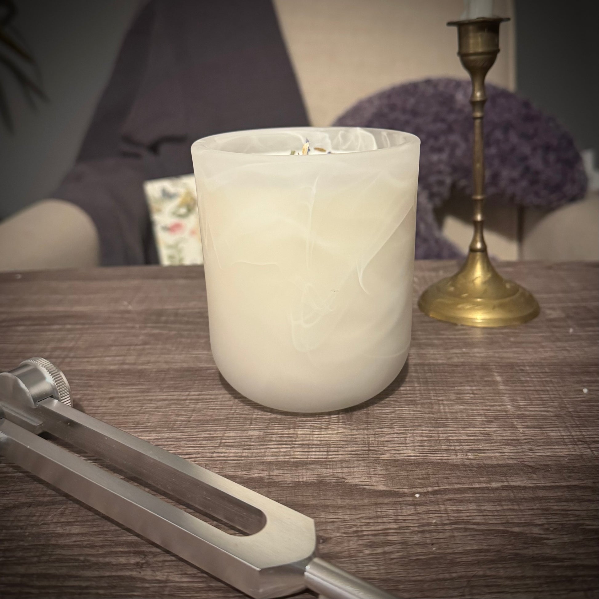 A white swirl glass candle sits on a table behind a tuning fork and in front of a brass candlestick