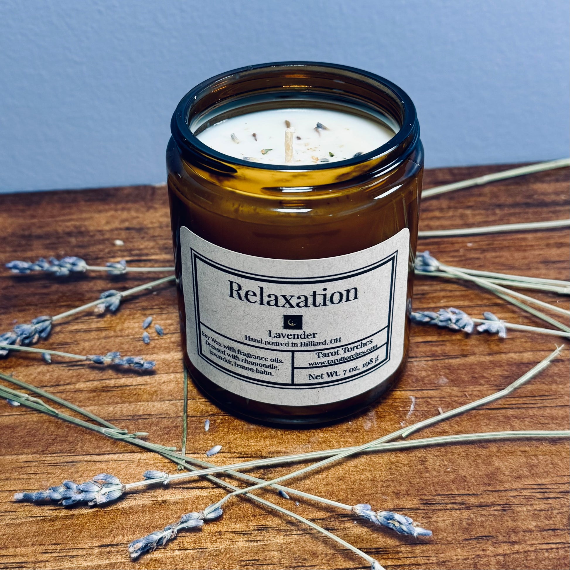 A brown glass candle with a label that reads 'Relaxation' and is infused with lavender fragrance. The candle is placed on a wooden surface with some dried lavender sprigs around it.