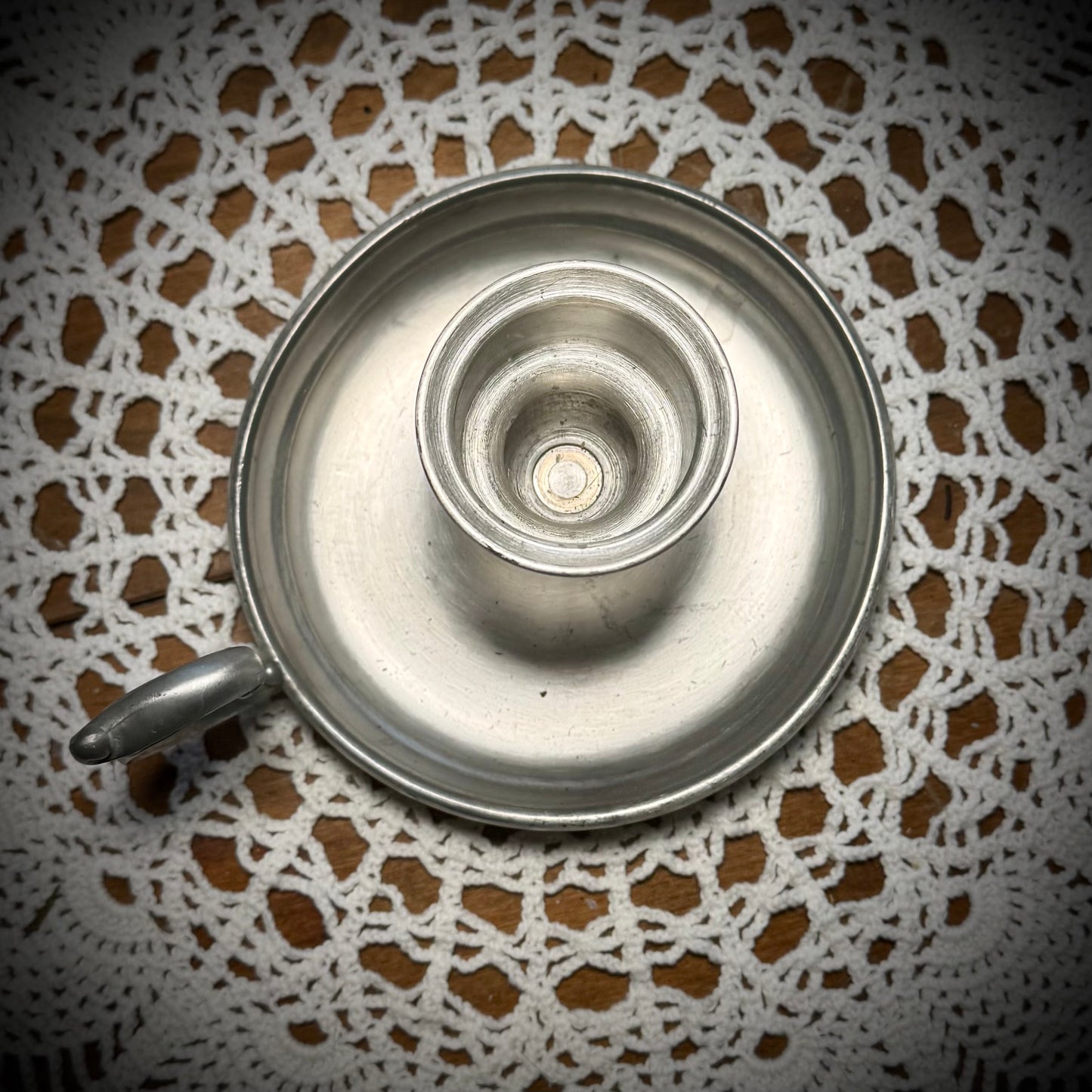 A vintage pewter candlestick with a handle, displayed on a table with a lace tablecloth, against a backdrop of a teapot and cups.