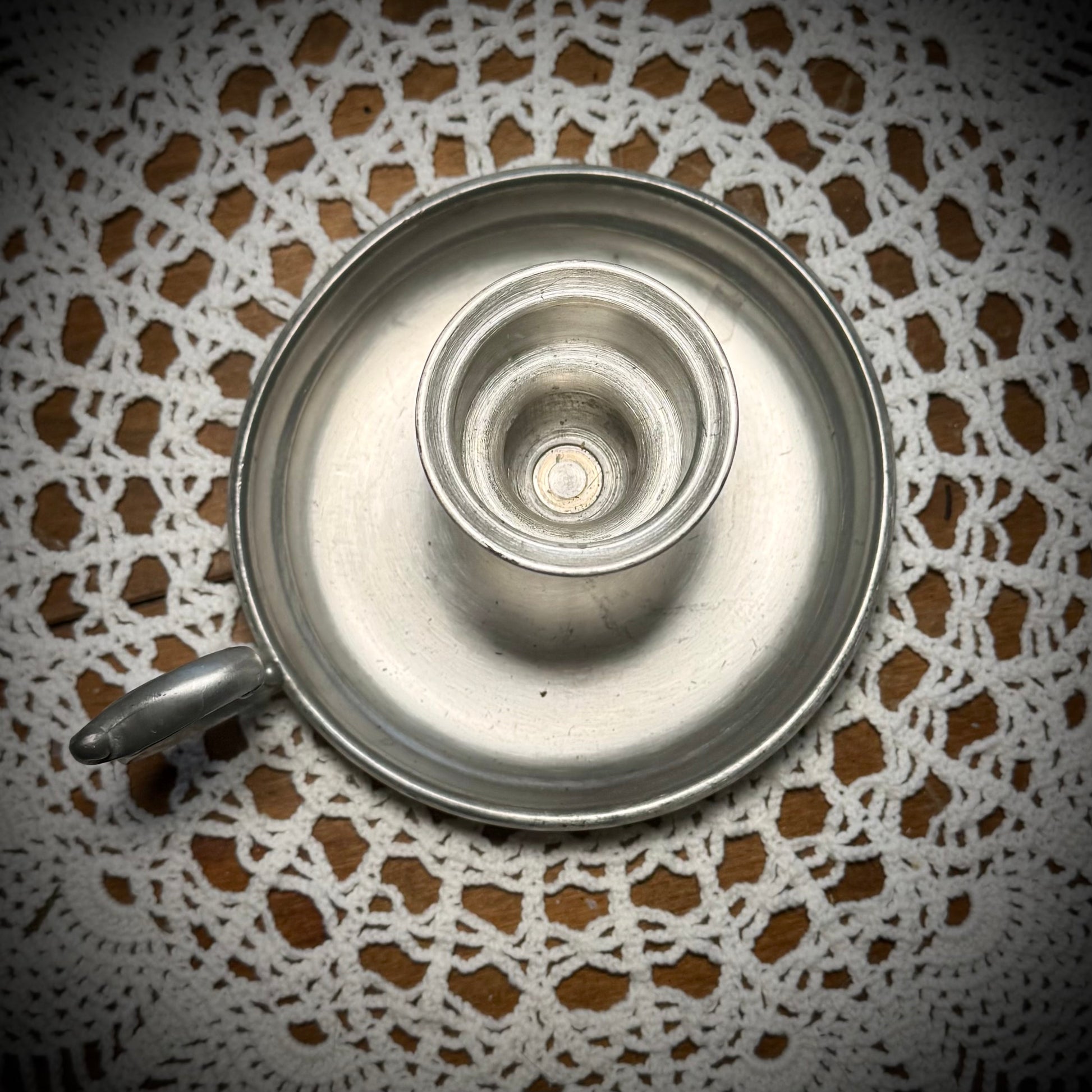 A vintage pewter candlestick with a handle, displayed on a table with a lace tablecloth, against a backdrop of a teapot and cups.