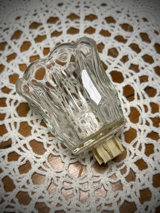 A clear glass votive candle holder with a textured design and a wooden peg at the bottom, placed on a lace tablecloth.