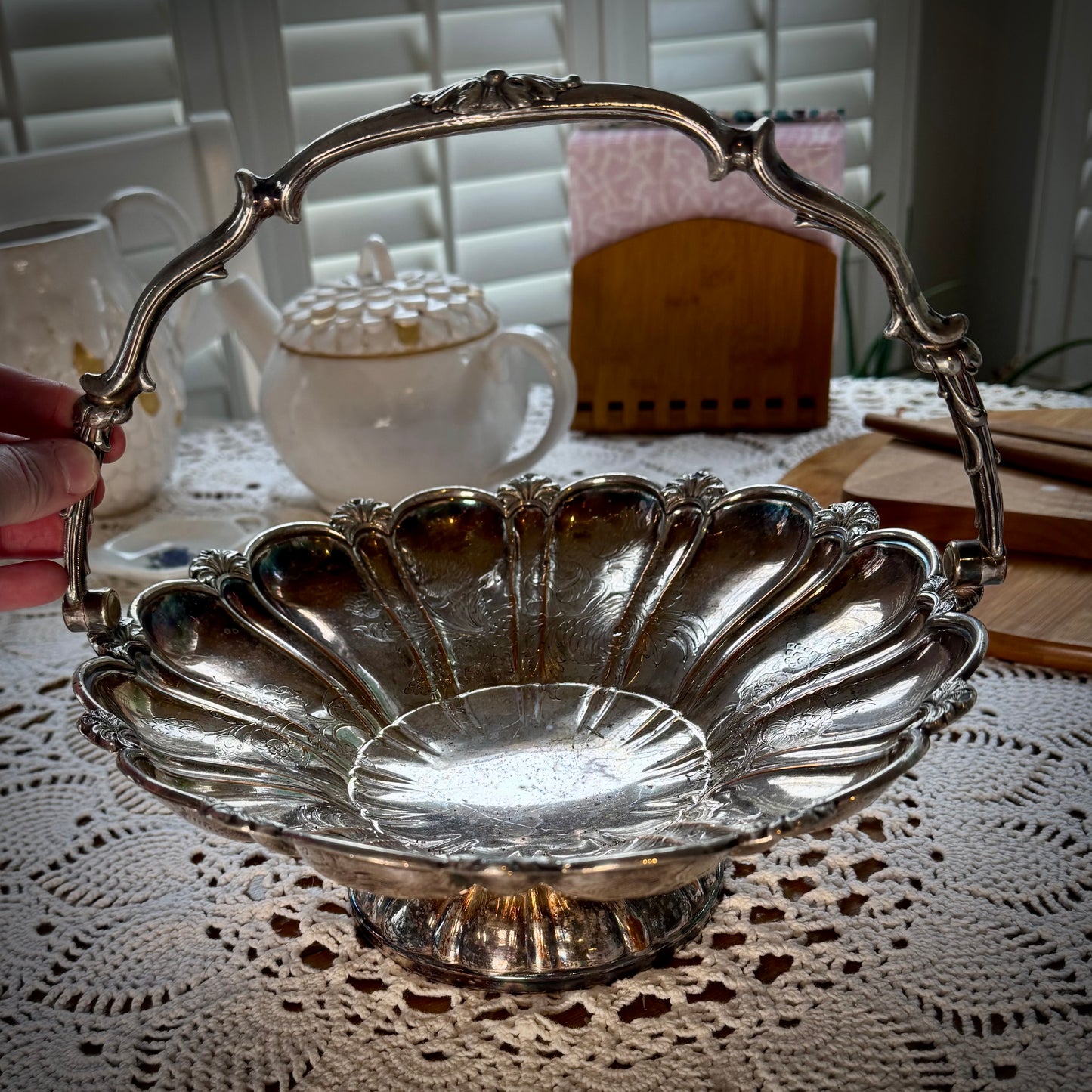 A silver plate basket with a handle, placed on a table with a lace tablecloth, displaying a reflective surface.