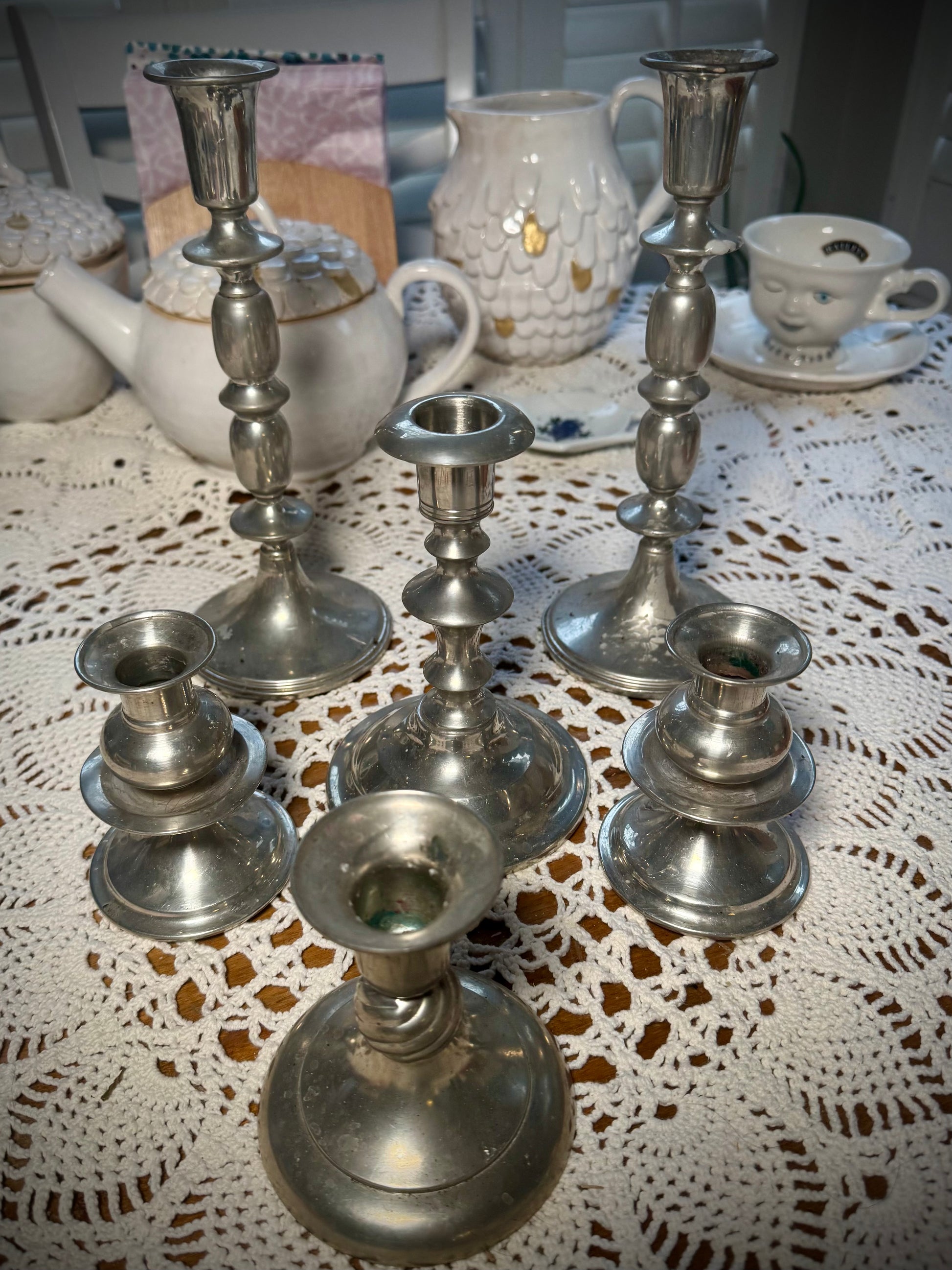 A collection of vintage pewter candlesticks in various sizes displayed on a table with a lace tablecloth.