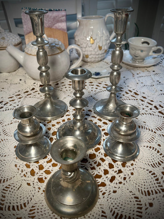 A collection of vintage pewter candlesticks in various sizes displayed on a table with a lace tablecloth.