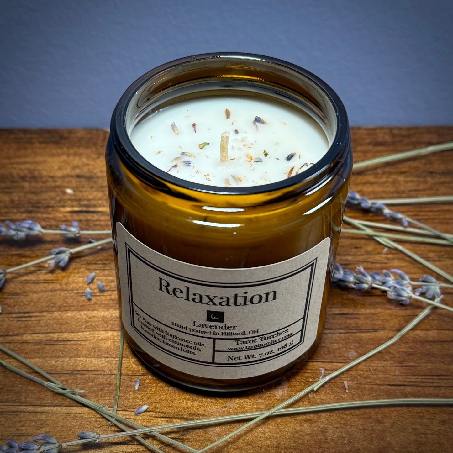 A brown glass candle with a label that reads 'Relaxation' and is infused with lavender fragrance. The candle is placed on a wooden surface with some dried lavender sprigs around it.