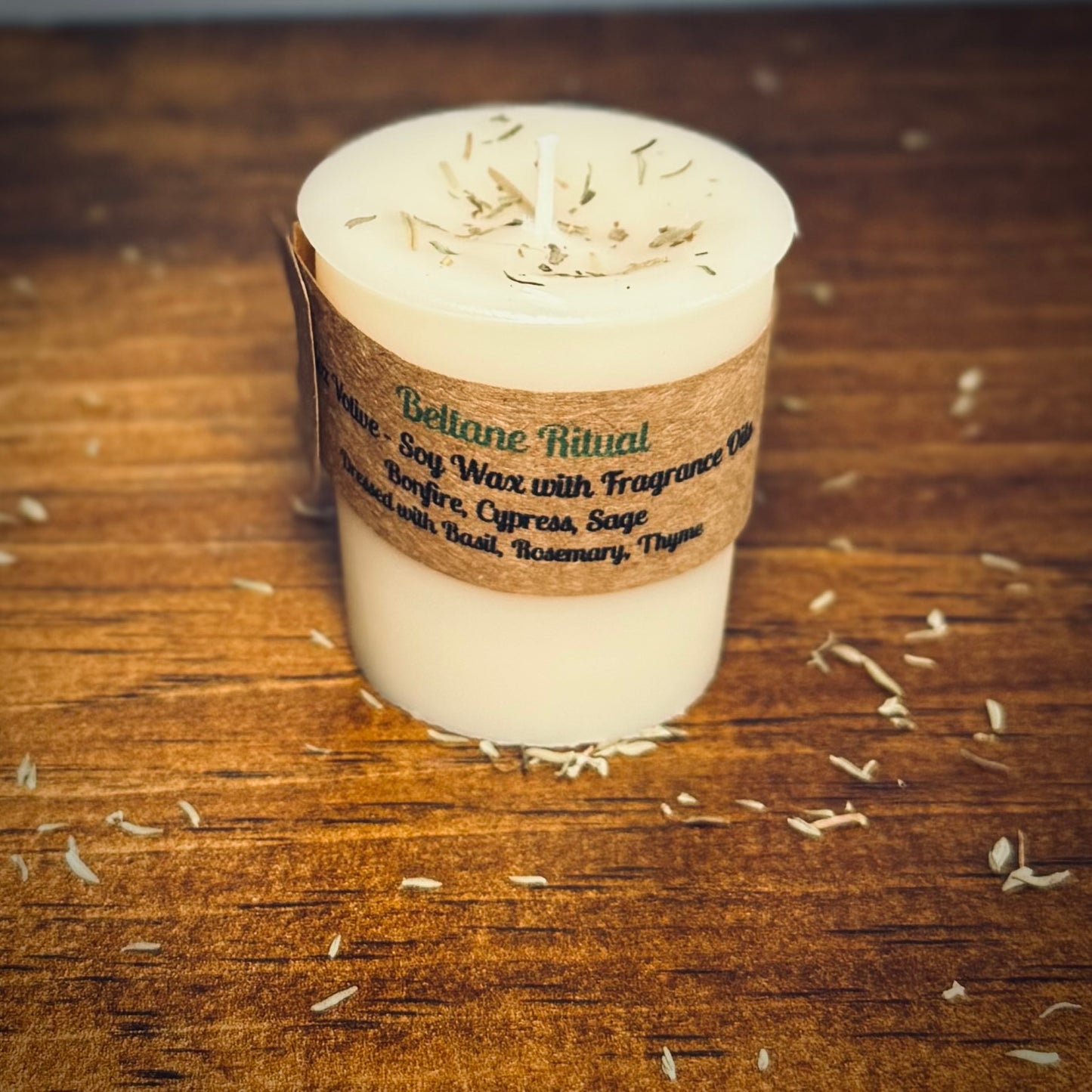 A white votive candle with a label reading "Beltane Ritual" sits on a wood surface surrounded by herbs.