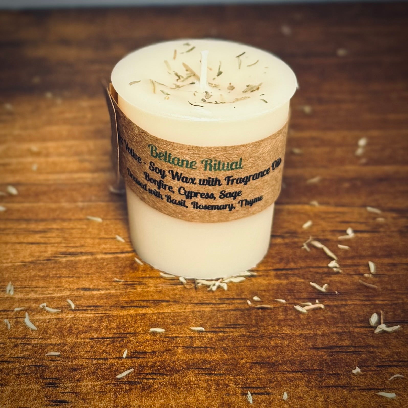 A white votive candle with a label reading "Beltane Ritual" sits on a wood surface surrounded by herbs.