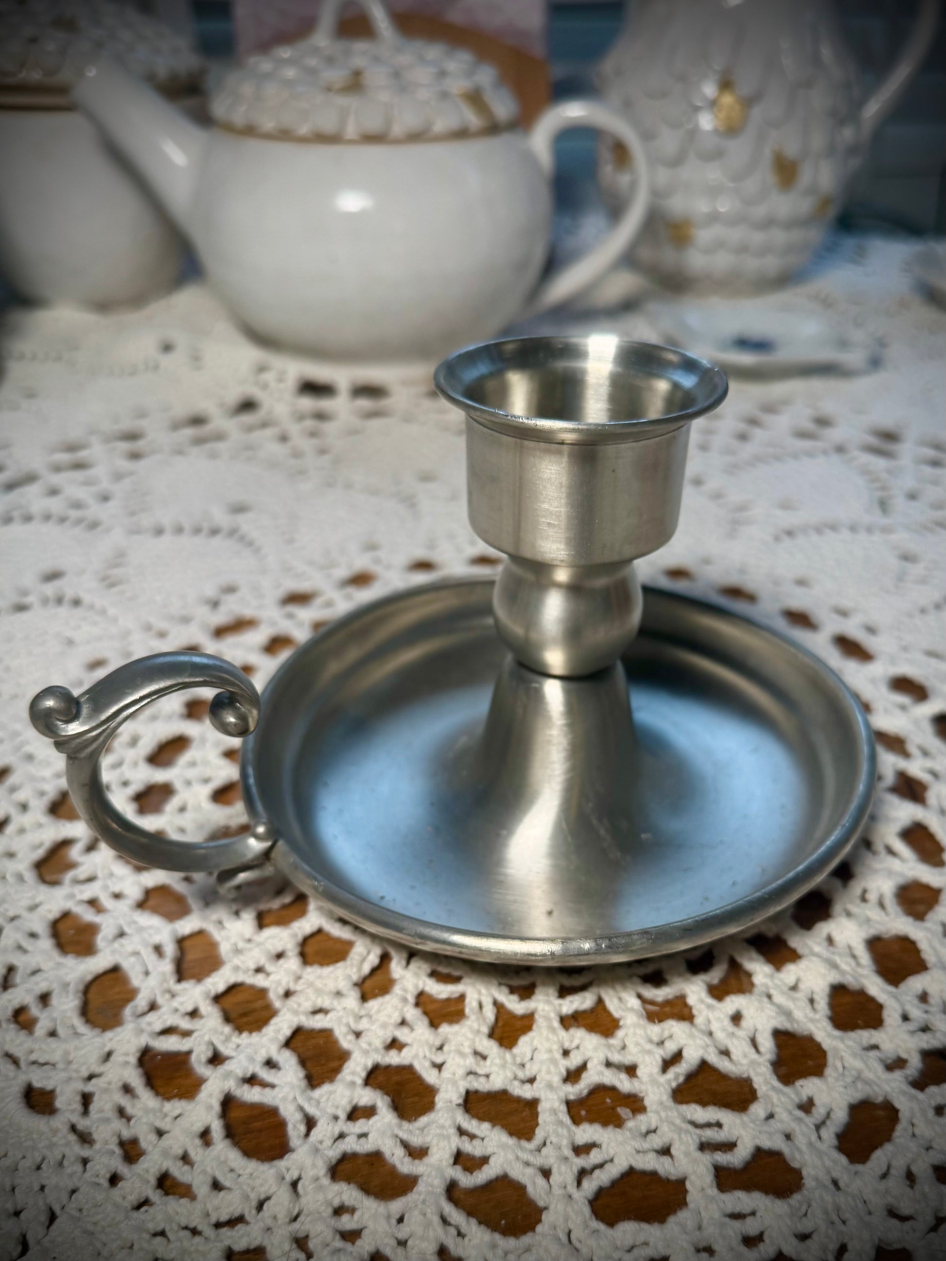 A vintage pewter candlestick with a handle, displayed on a table with a lace tablecloth, against a backdrop of a teapot and cups.