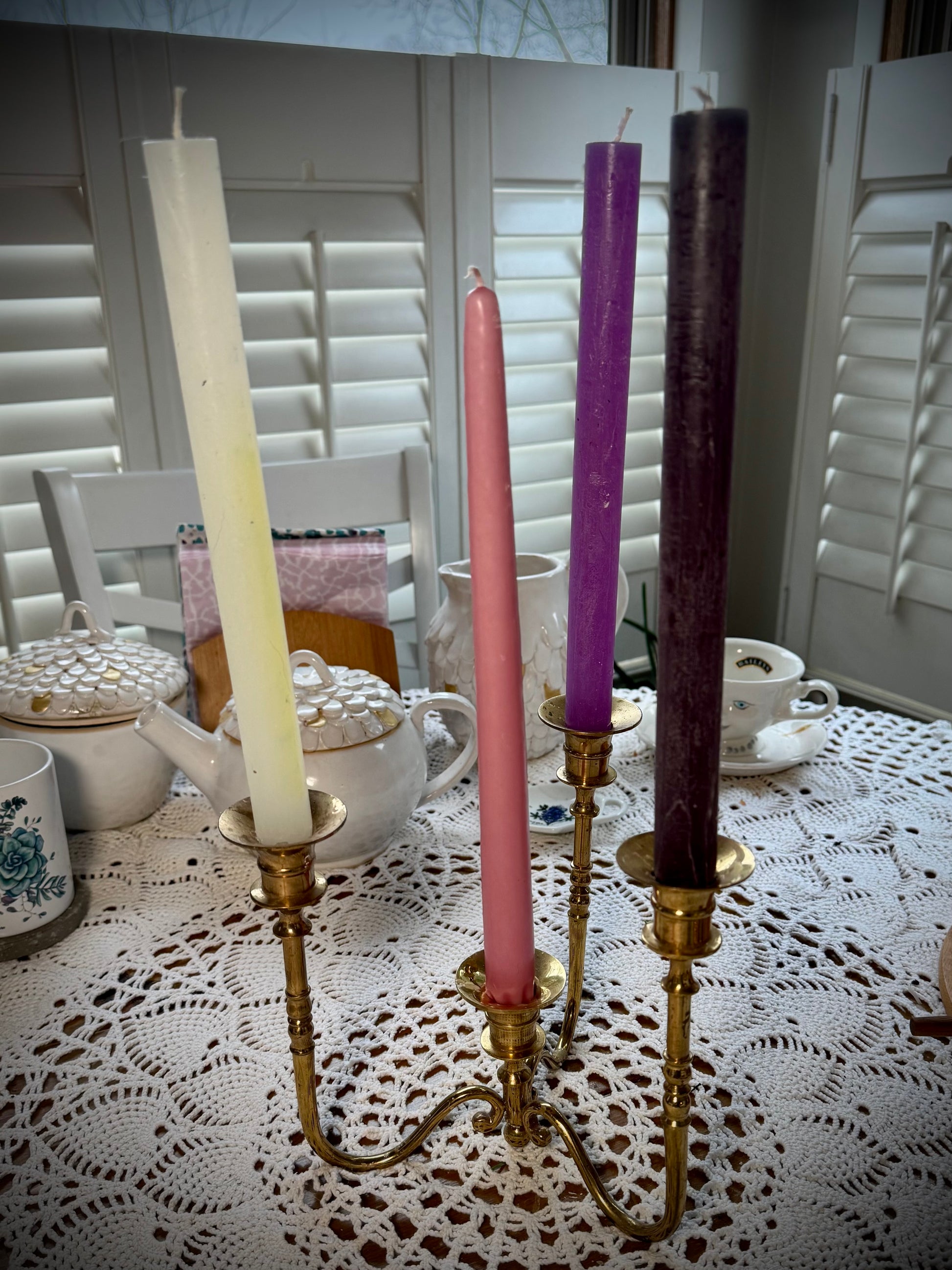 A vintage brass four-arm candelabra placed on a table with a lace tablecloth, holding four different color taper candles.