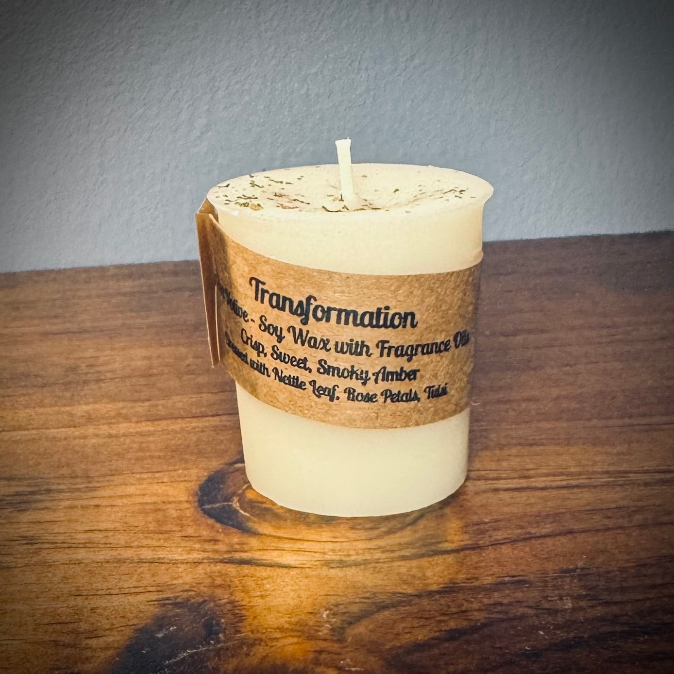A white votive candle with a kraft label reading "Transformation" sits on a wooden surface