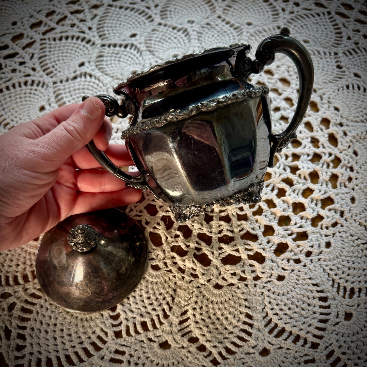 Vintage silver plate sugar bowl with intricate designs, showing signs of wear, being held by a hand