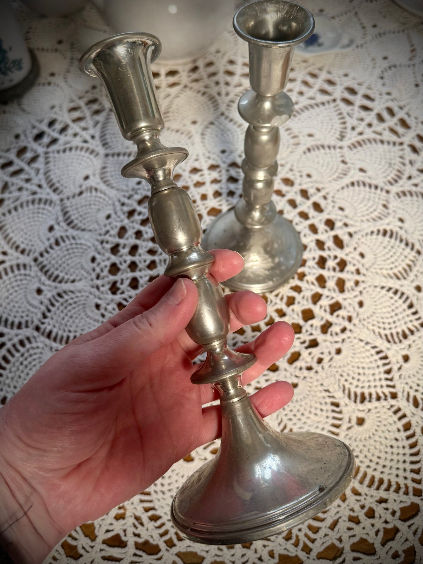 A tall vintage pewter candlestick being held with a similar candlestick in the background