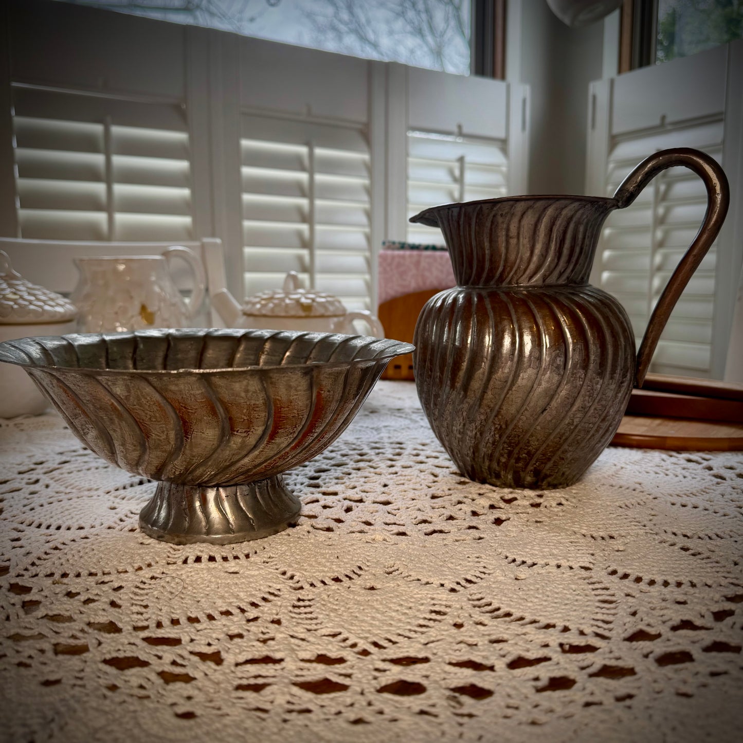 An antique swirl tinned copper pitcher and bowl placed on a table with a lace tablecloth.