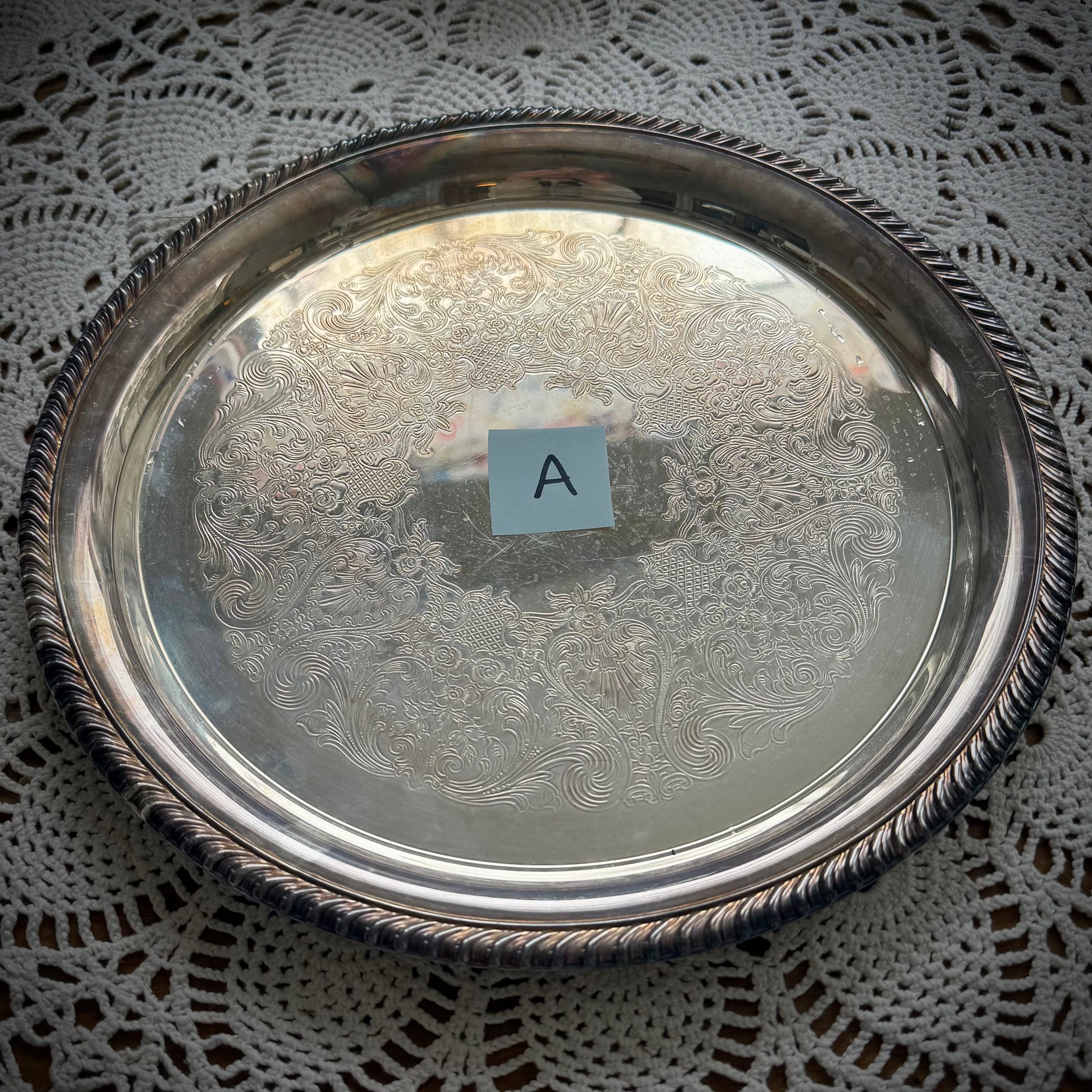 A silverplate round offering tray is displayed on a white lace surface. The tray has a post-in with the letter "A"