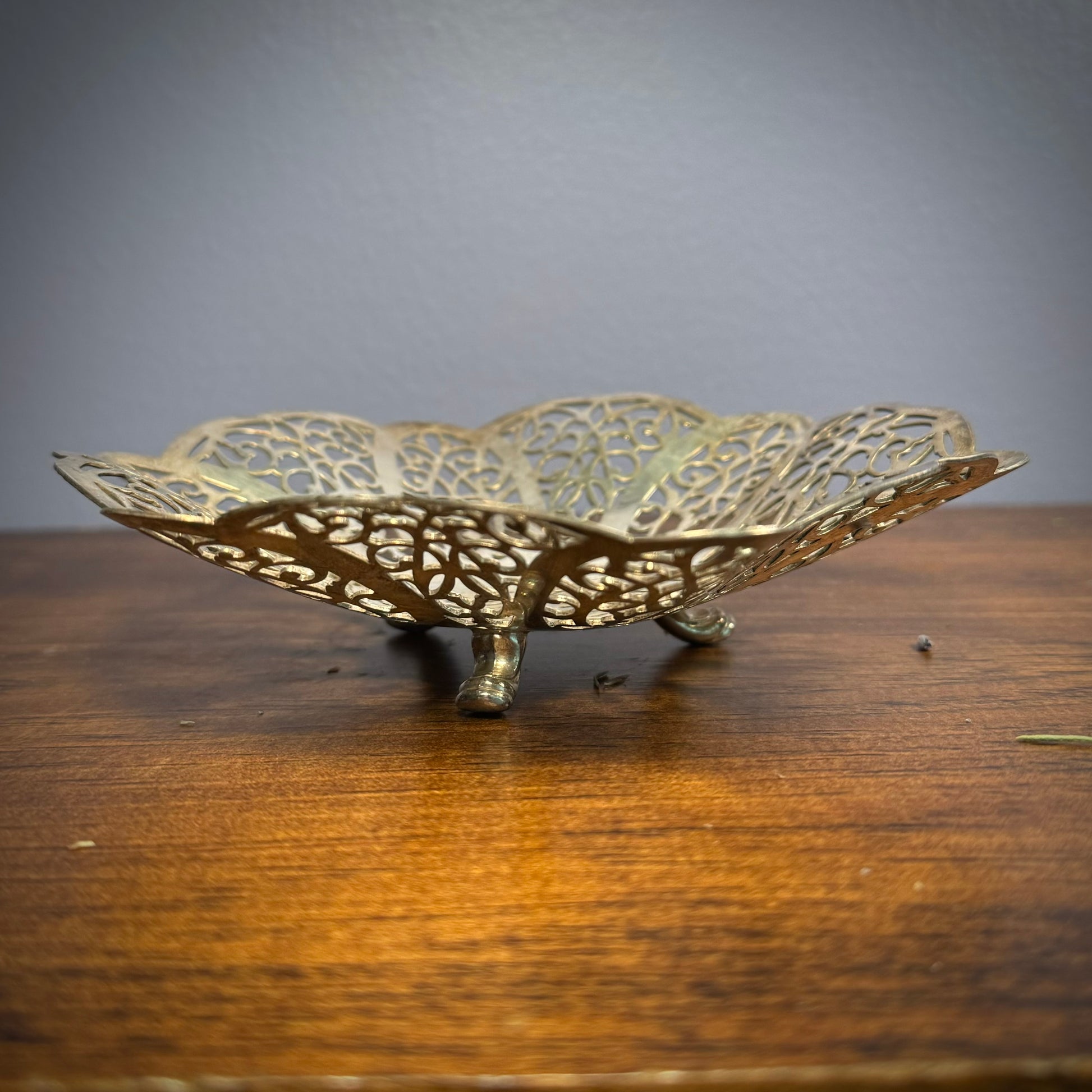 A vintage silver ritual plate with intricate floral lace patterns and three small feet on a wooden surface.