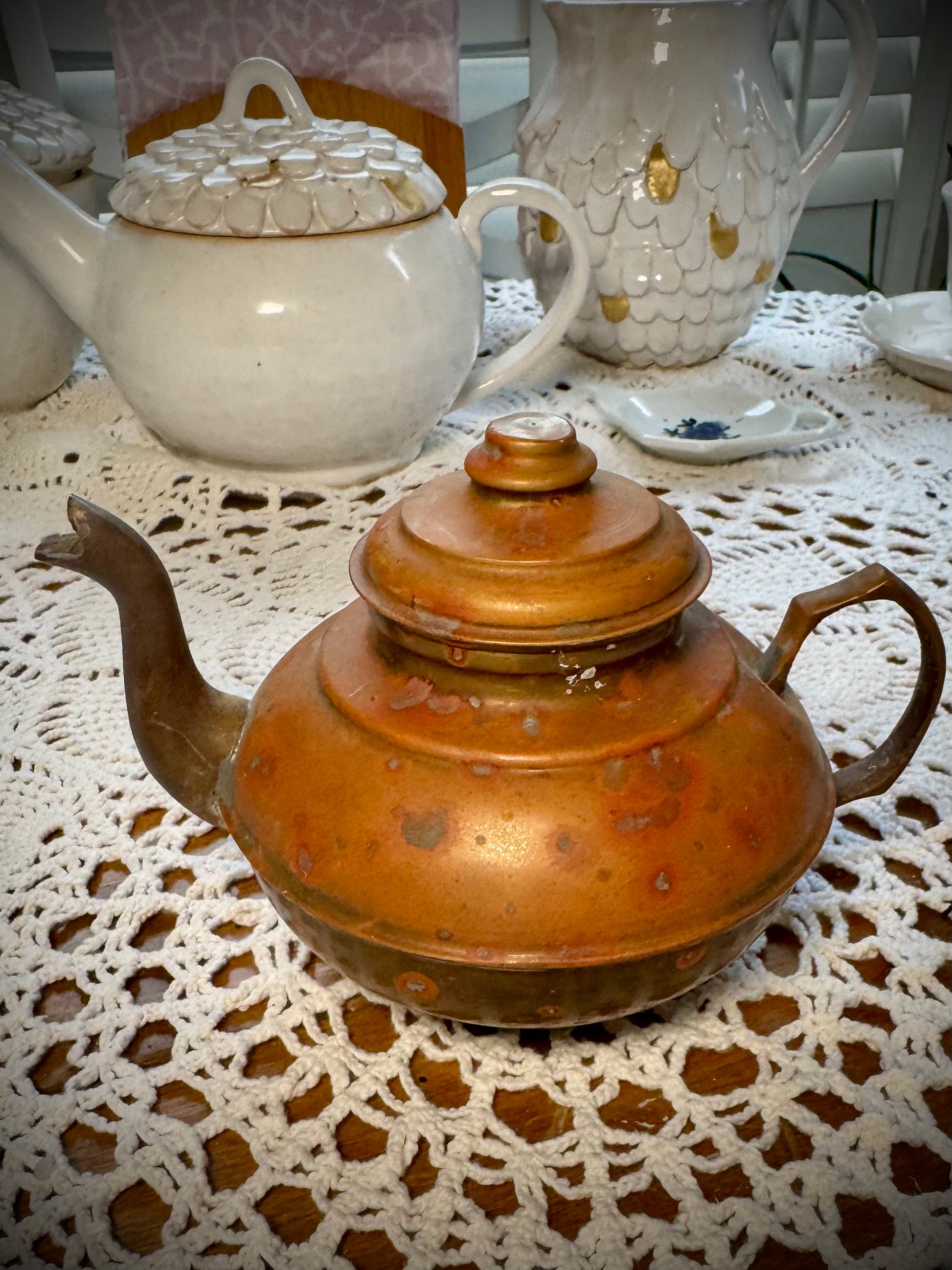 A vintage copper teapot with a Middle Eastern style design, placed on a table with a white lace tablecloth.