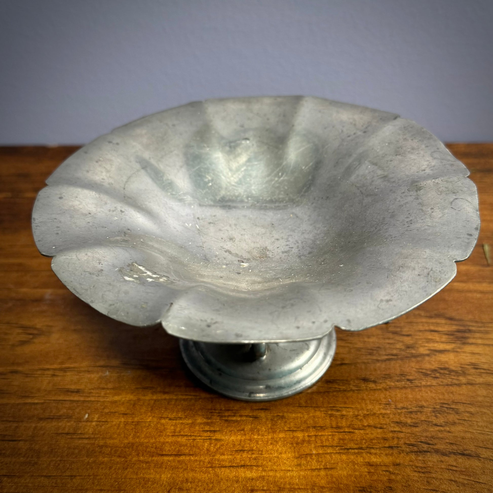 A silver pewter bowl with three detailed feet attached to a round base, placed on a wooden surface with a plain blue background.