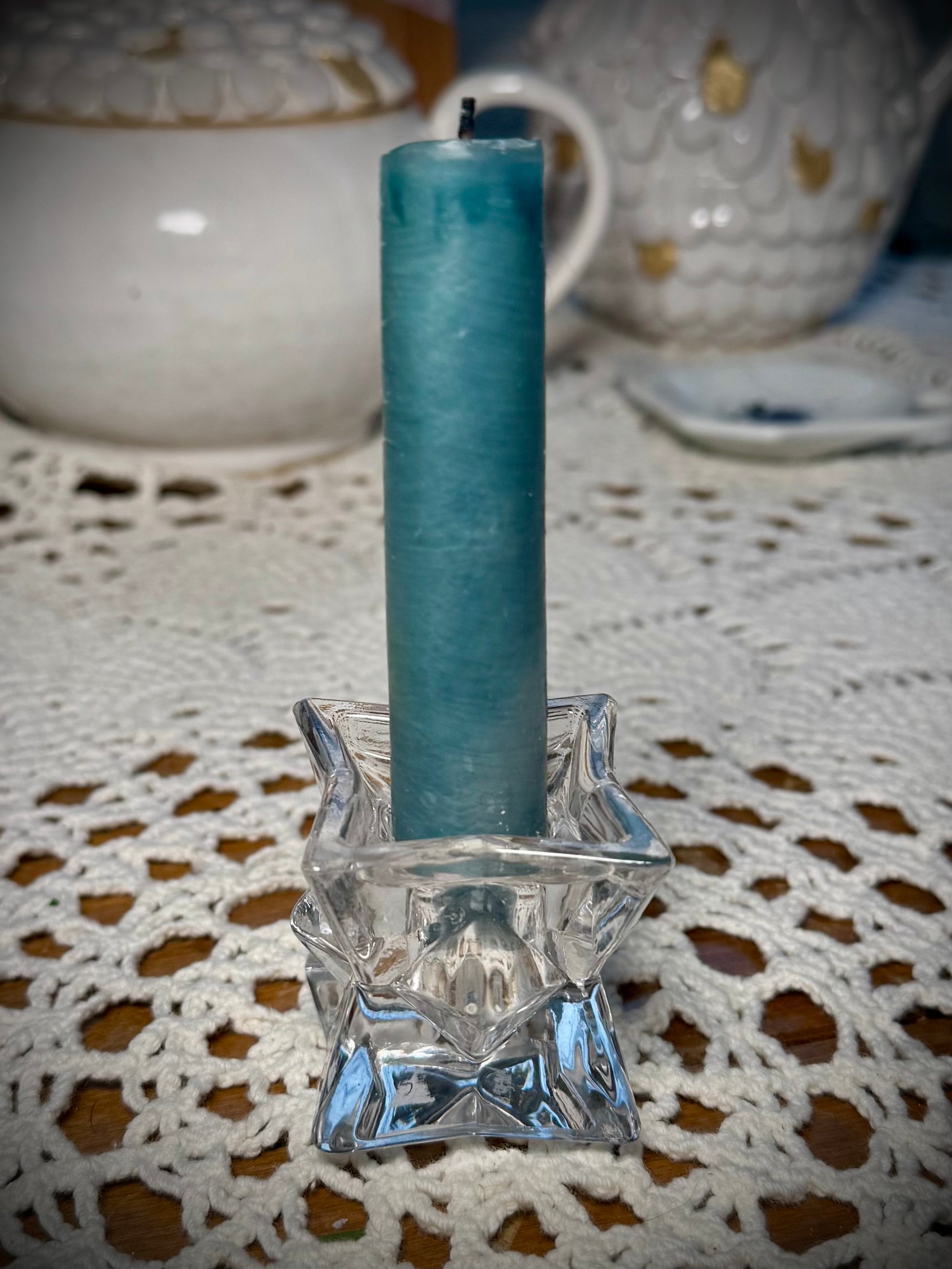 A small glass geometric candle holder with a blue taper candle in it on a table with the white lace tablecloth.