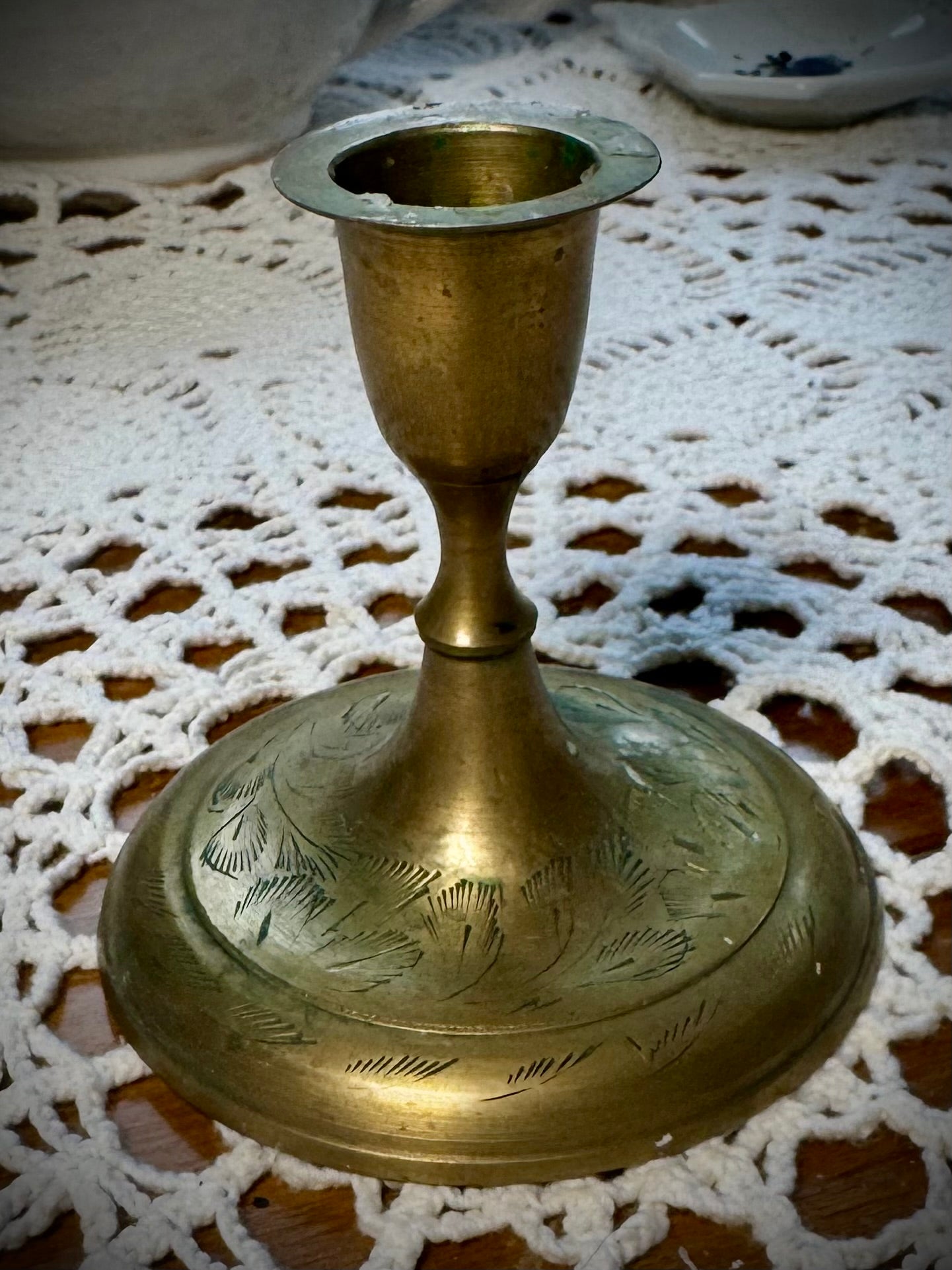 A vintage brass taper candle holder with etching on the base, displayed on a lace tablecloth.